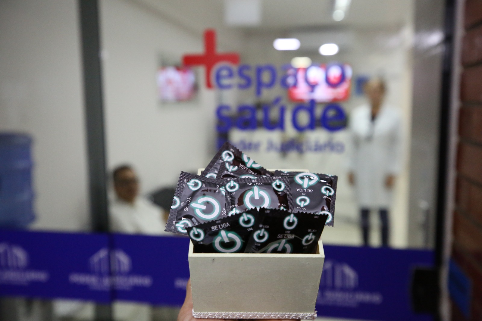 Fotografia colorida de um recipiente com preservativos na frente de um vidro com os dizeres Espaço Saúde