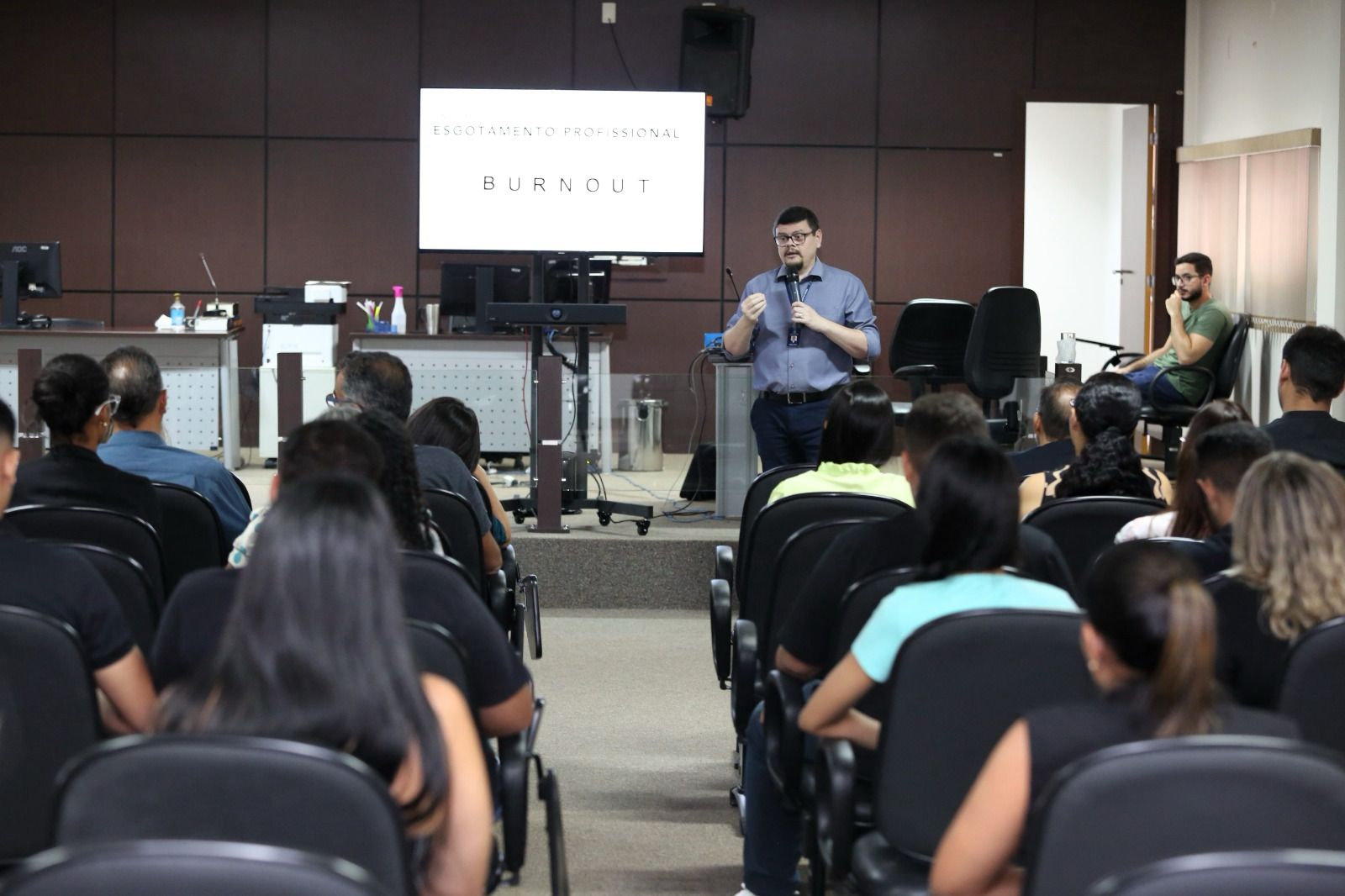 Fotografia colorida do psiquiatra do NAPsi, Wordney Carvalho Camarço, palestrando ao lado de uma televisão escrita burnout para um grupo de pessoas sentados em sua frente