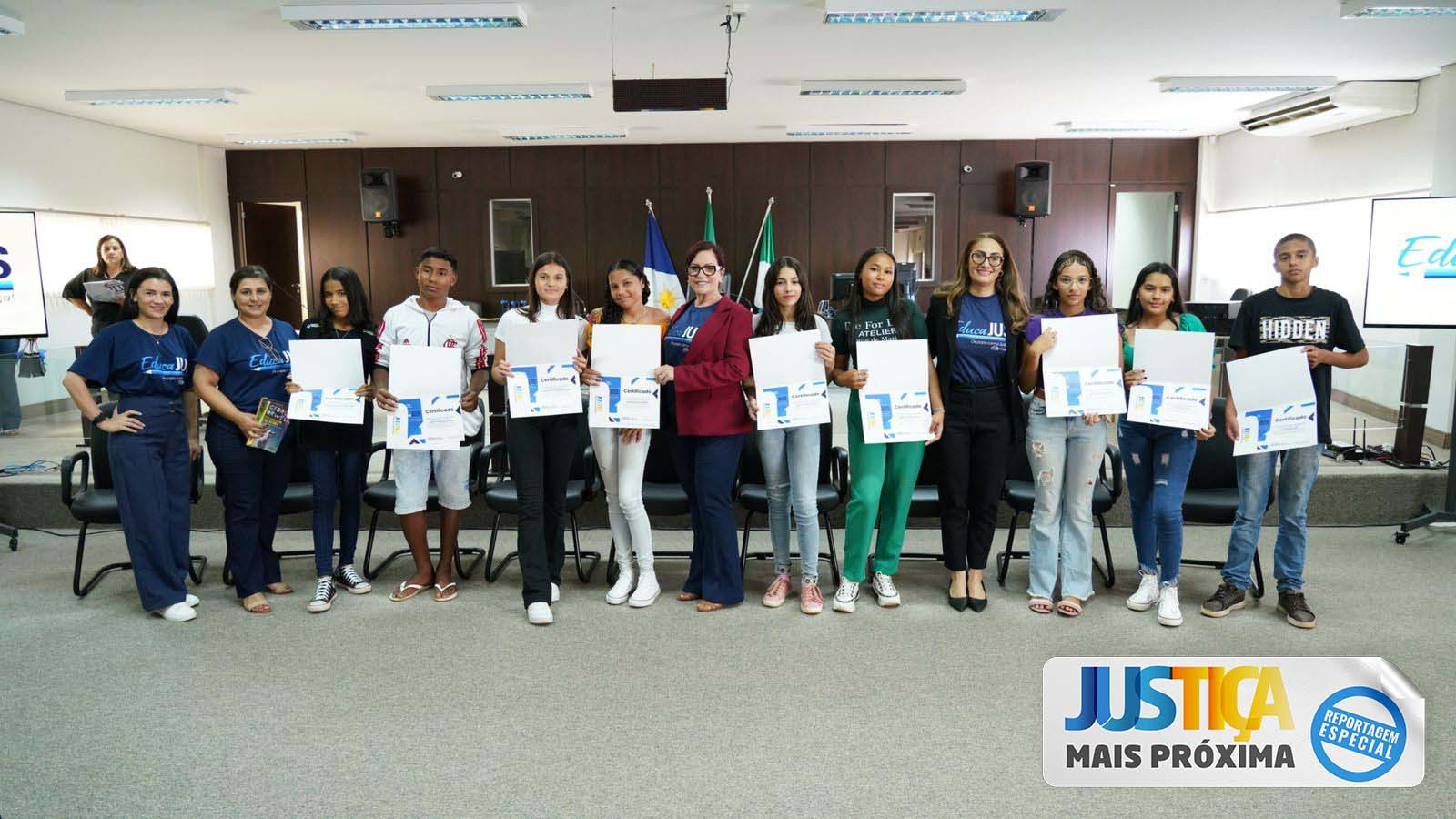 presidente do TJTO, de terno vermelho e calça preta, ao lado da juíza Rosa Maria Rossi, intergrante do projeto e estudantes premiados no projeto Educajus; na parte inferior à direita, um selo com o nome Justiça Mais Próxima - Reportagem Especial