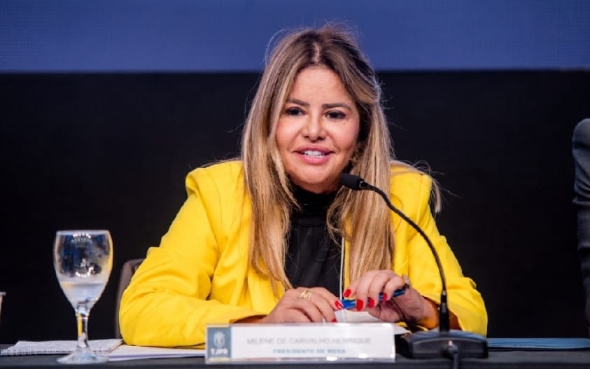 Na fotografia tem apenas uma mulher loira, de blazer amarelo e por baixo uma blusa preta, ela fala ao microfone com um leve sorriso e ao lado dela na mesa tem uma taça com água