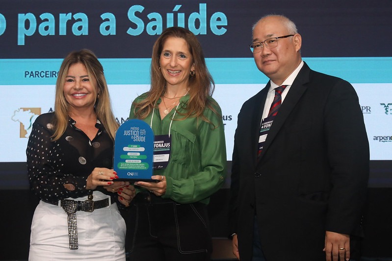 Na fotografia tem três pessoas posando para a foto, uma mulher branca e loira, de blusa preta, uma mulher um pouco mais alta, branca, de cabelos castanhos e blusa verde, ambas estão segurando o prêmio do CNJ e ao lado da mulher de blusa verde tem um homem de terno preto e gravata listrada, usando óculos de grau