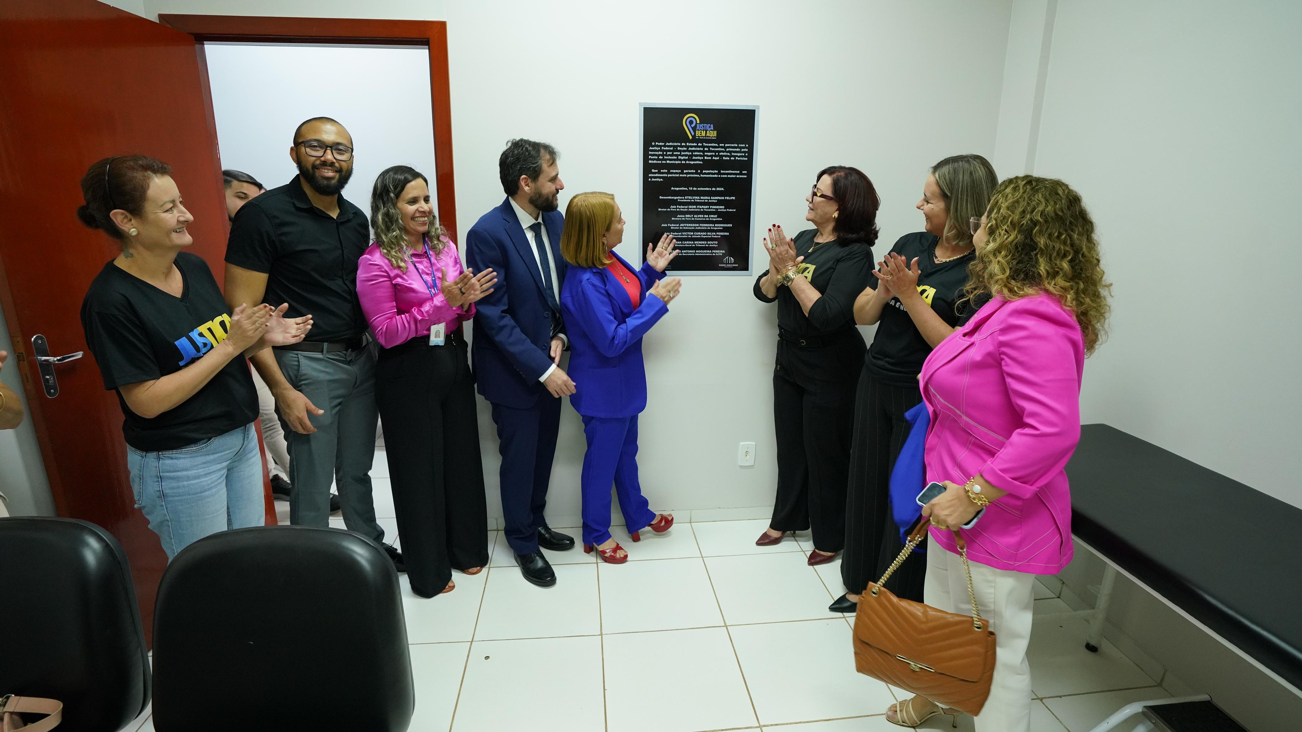 Grupo de pessoas admirando e aplaudindo uma placa em uma sala de paredes brancas.