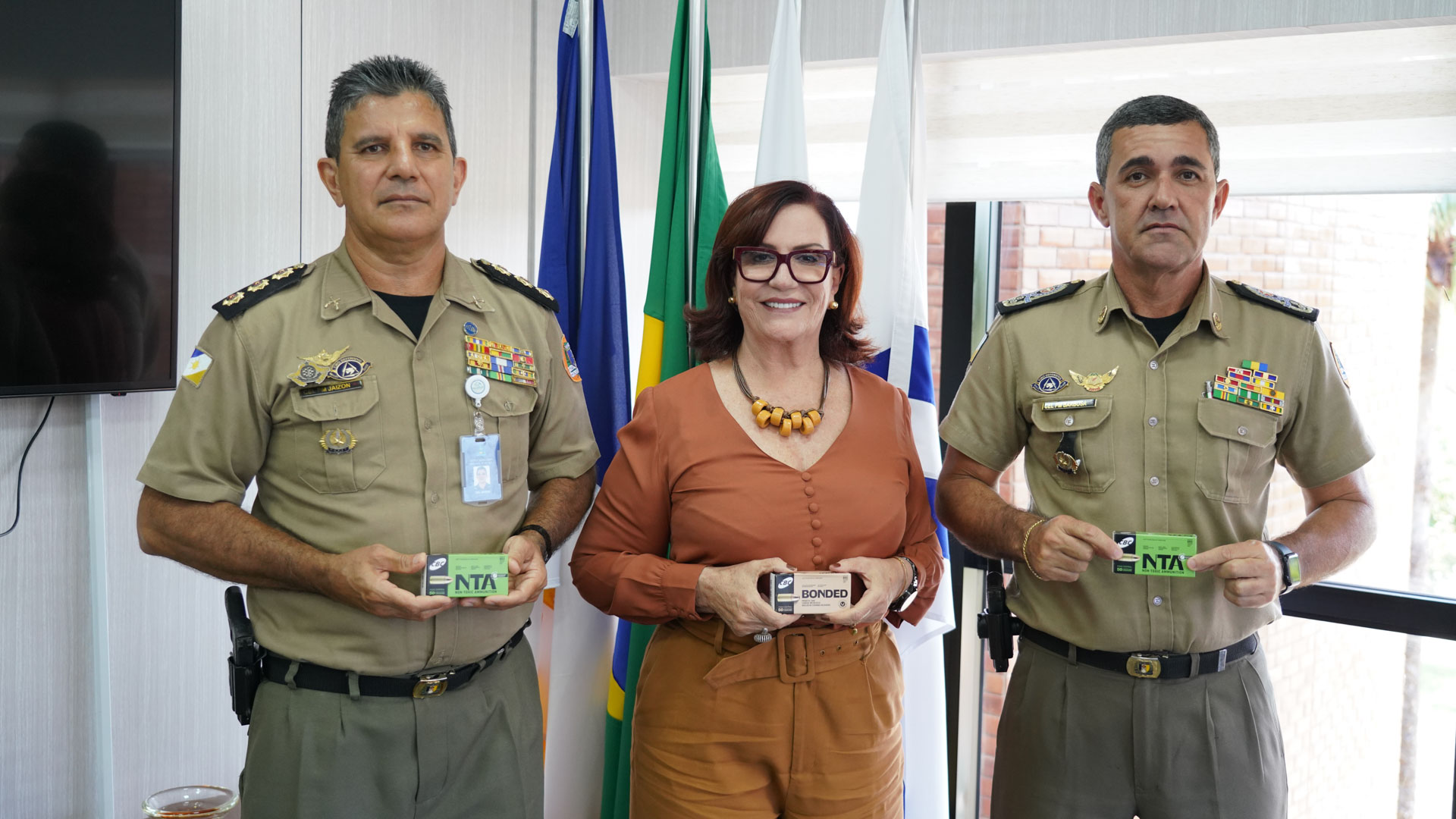 Foto mostra três pessoas; dois homens com fardas da PM em pé e uma mulher, a presidente do TJTO; todos estão segurando caixas com  munições