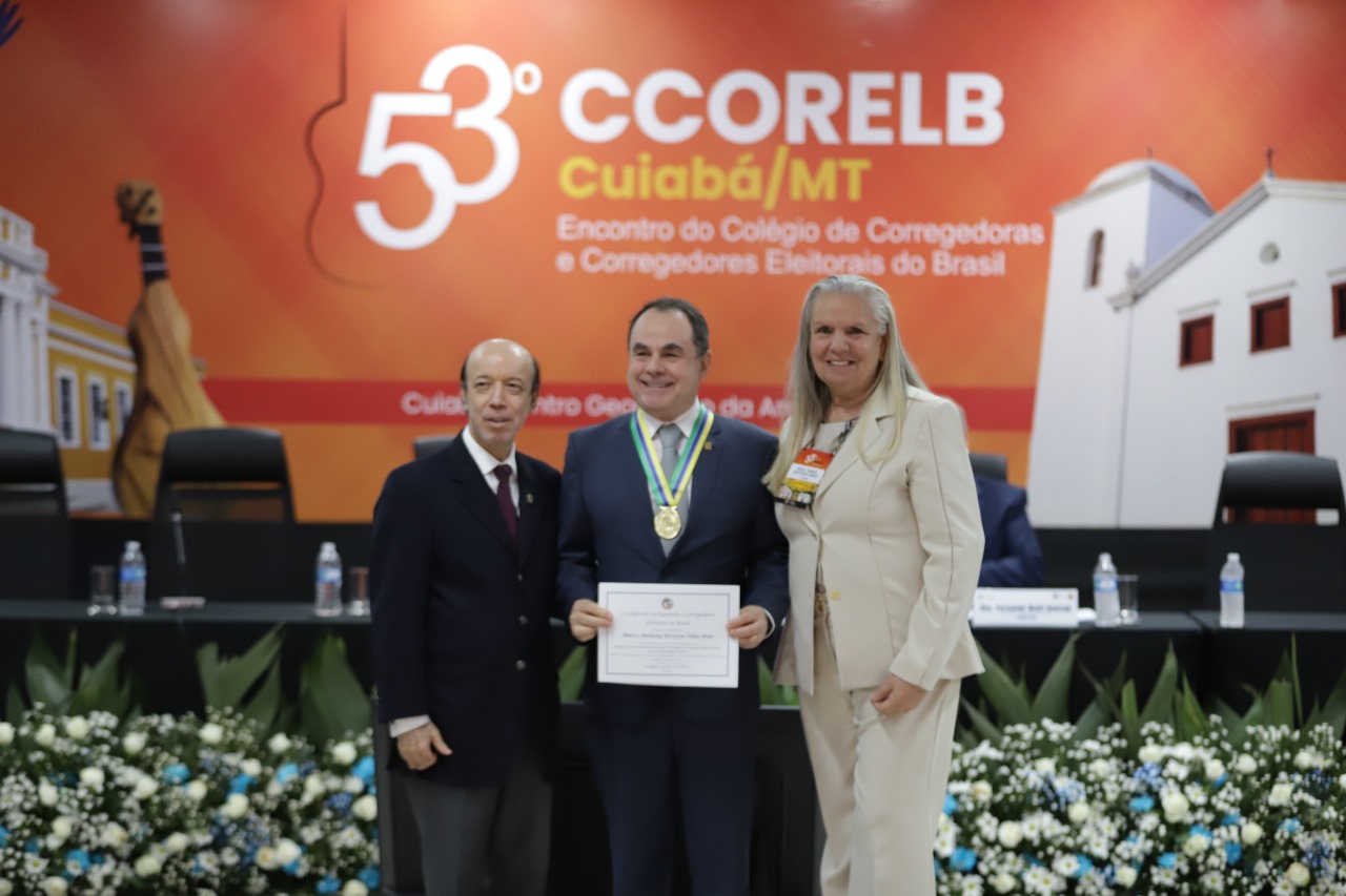 Homem pardo, de cabelos levemente grisalhos, de terno azul escuro, com medalha no pescoço, segurando diploma entre homem pardo, calvo, de terno preto e mulher branca de terno claro.
