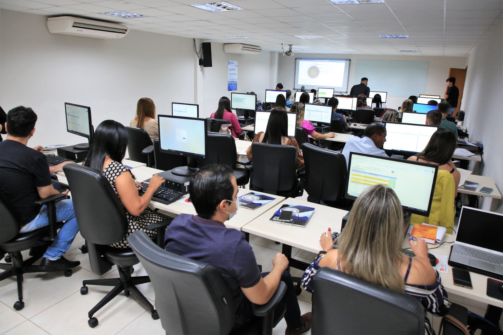 Foto com várias pessoas sentadas em mesas e à frente computadores; todos estão em curso