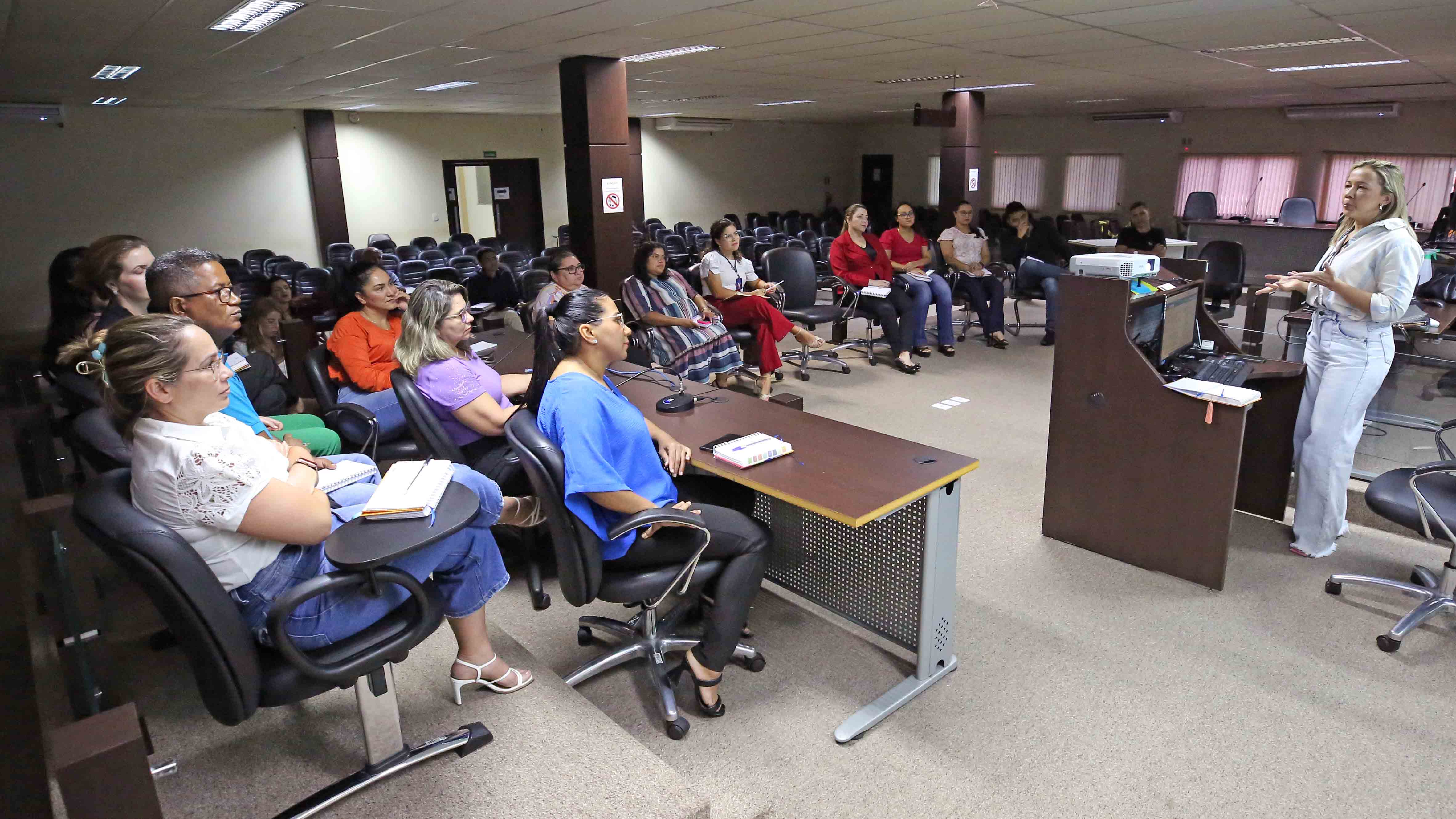 Homens e mulheres sentados durante treinamento ministrado por mulher que está em pé, de calça jeans clara, camisa branca e cabelos loiros soltos