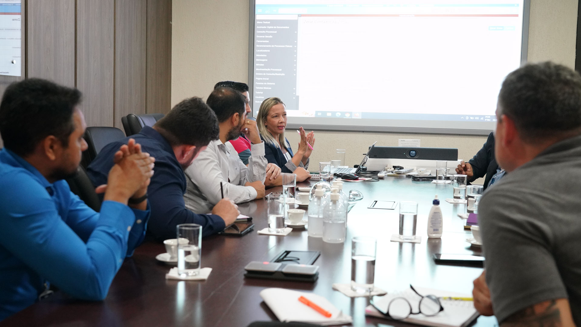Homens e mulheres sentados em torno de uma mesa de reunião de tampo de madeira marrom escuro, com parede revestida com o mesmo material 