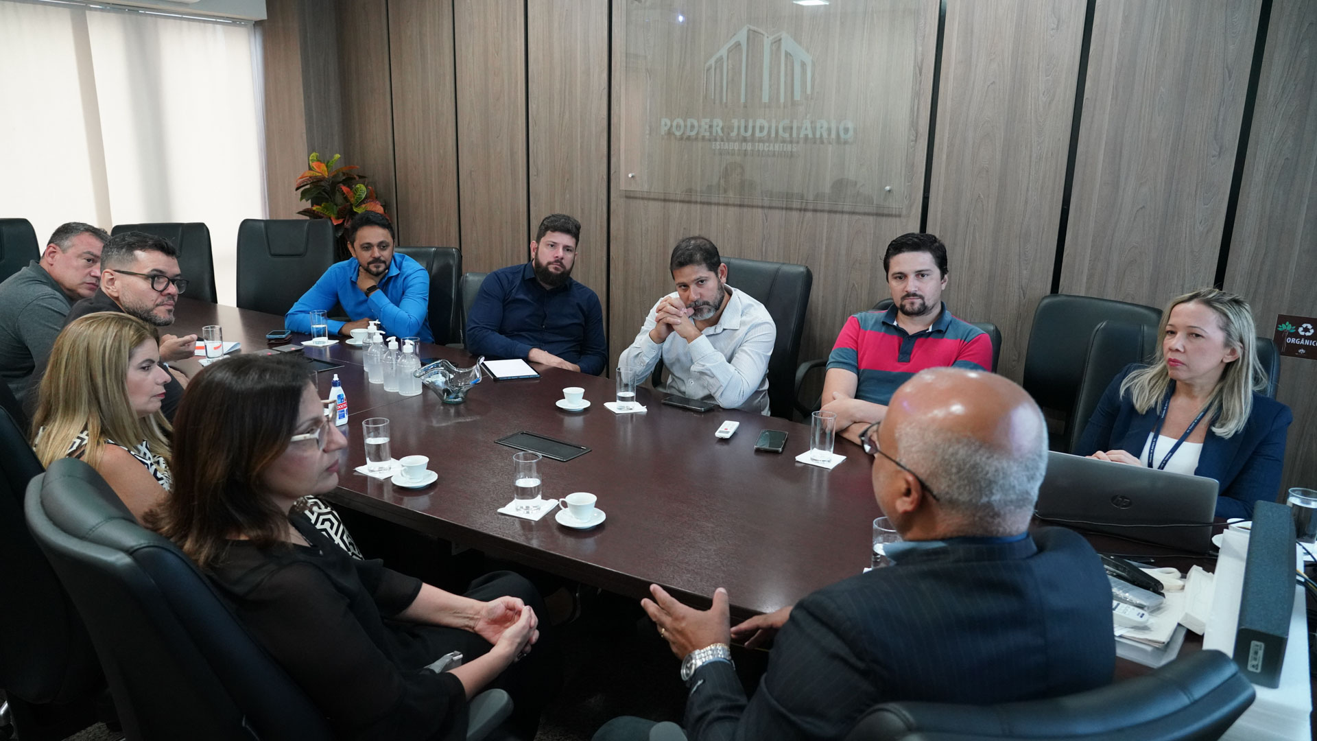 Homens e mulheres sentados em torno de uma mesa de reunião de tampo de madeira marrom escuro, com parede revestida com o mesmo material 