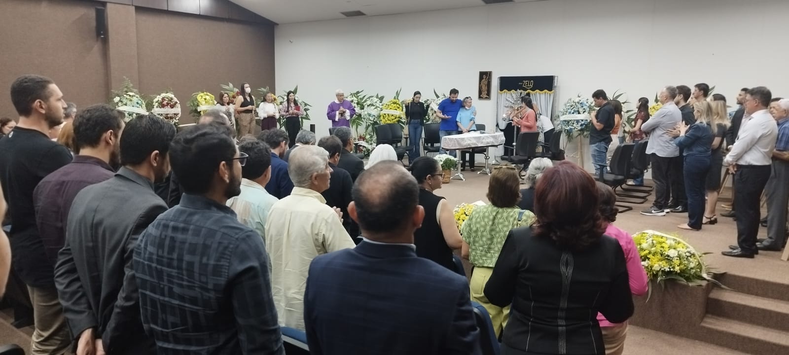 Homens e mulheres em pé durante velório de juiz que acontece no Fórum, uma sala com uma parede branca e outra marrom