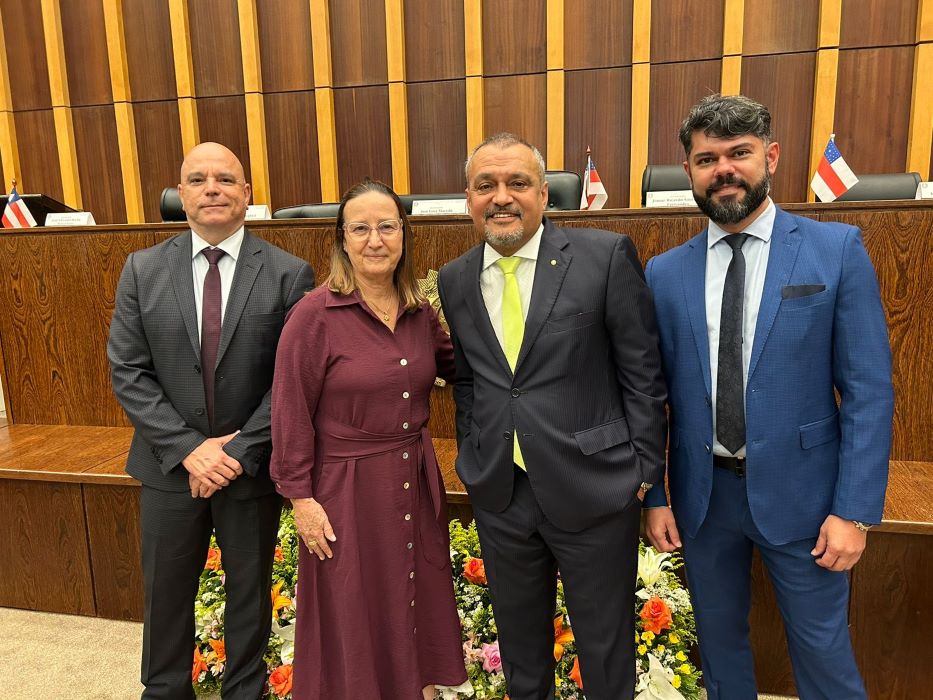 Fotografia colorida onde quatro corregedores. três homens e uma mulher, os homens de terno e a mulher com um vestido roxo vinho, posam para a foto em frente a uma mesa palanque de madeira marrom