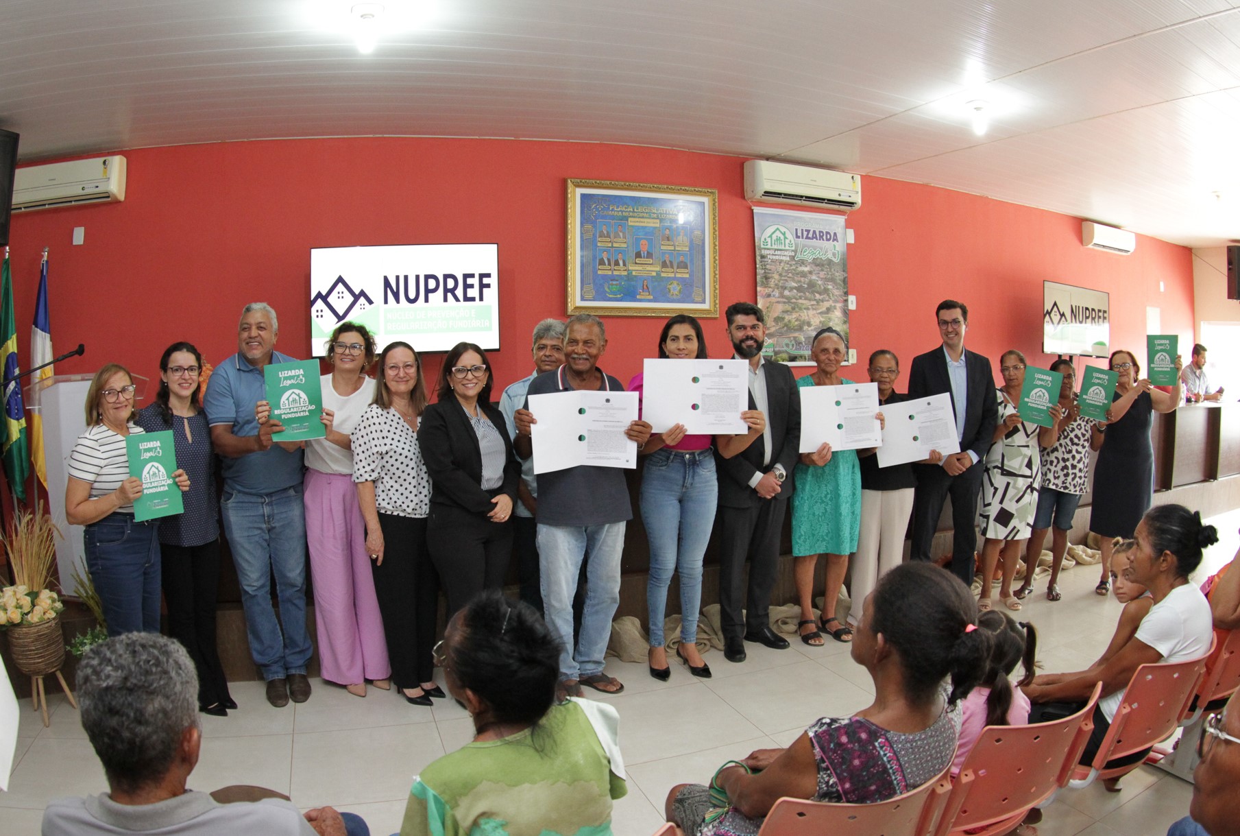 Fotografia colorida dos funcionários do judiciário juntamente com os moradores de Lizarda, em torno de 16 pessoas, segurando os documentos de regularização fundiária recebidos. Eles estão todos de pé posando para a foto em frente a uma parede alaranjada
