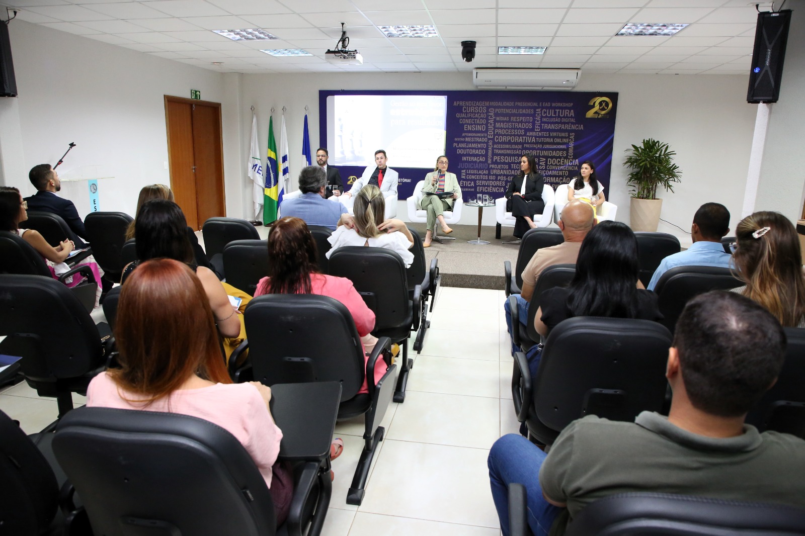 Fotografia colorida onde pessoas estão sentadas em uma plateia enquanto os professores (as) do curso Estratégias Para o Cumprimento de Metas ministram a aula sentados logo a afrente  