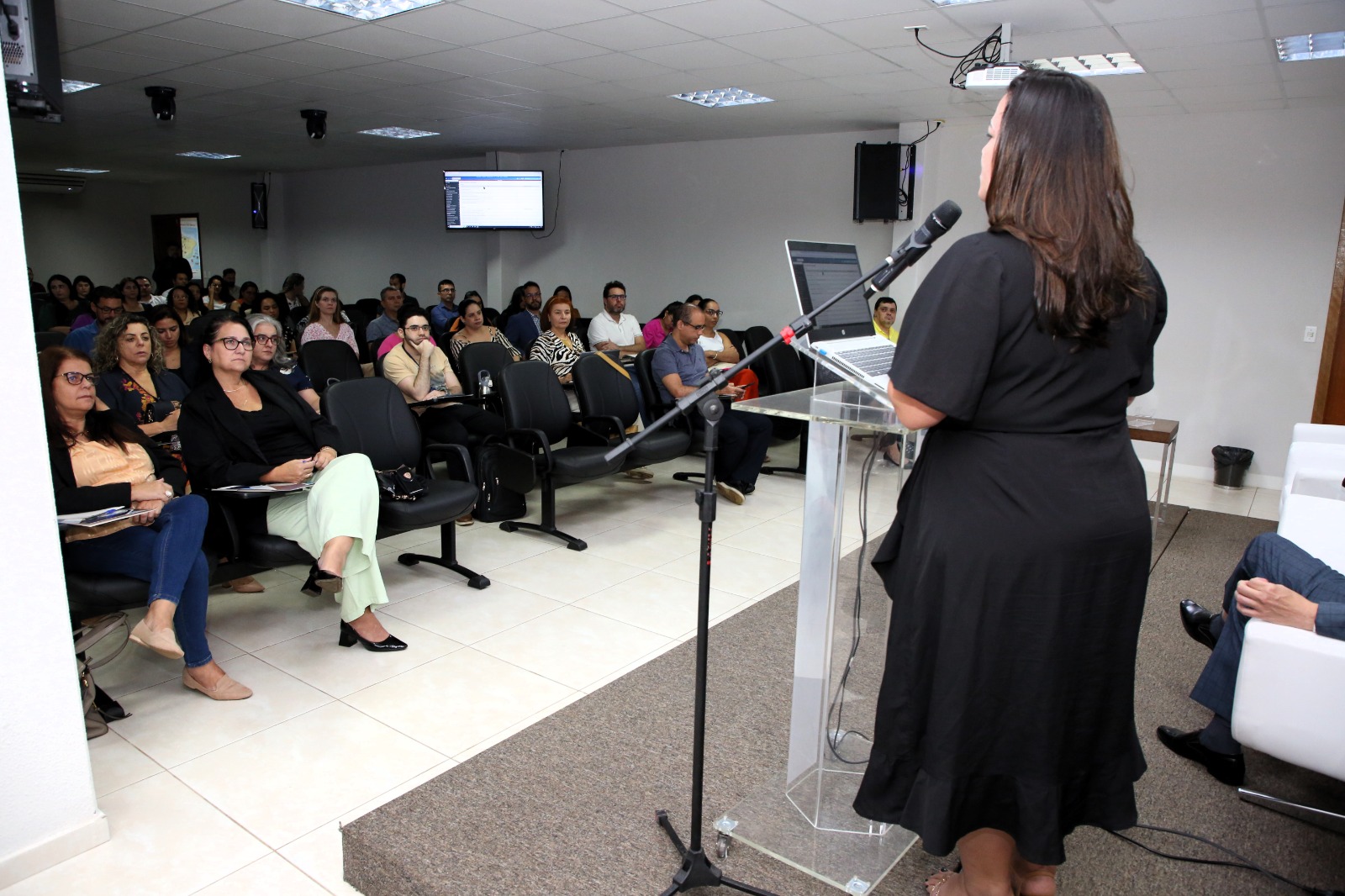 Fotografia colorida de uma mulher em pé em um palanque, usando vestido preto e com cabelo, ministrando a aula de Automatização de Localizadores no Sistema Eproc, enquanto os ministrados estão sentados na plateia assistindo a  aula 