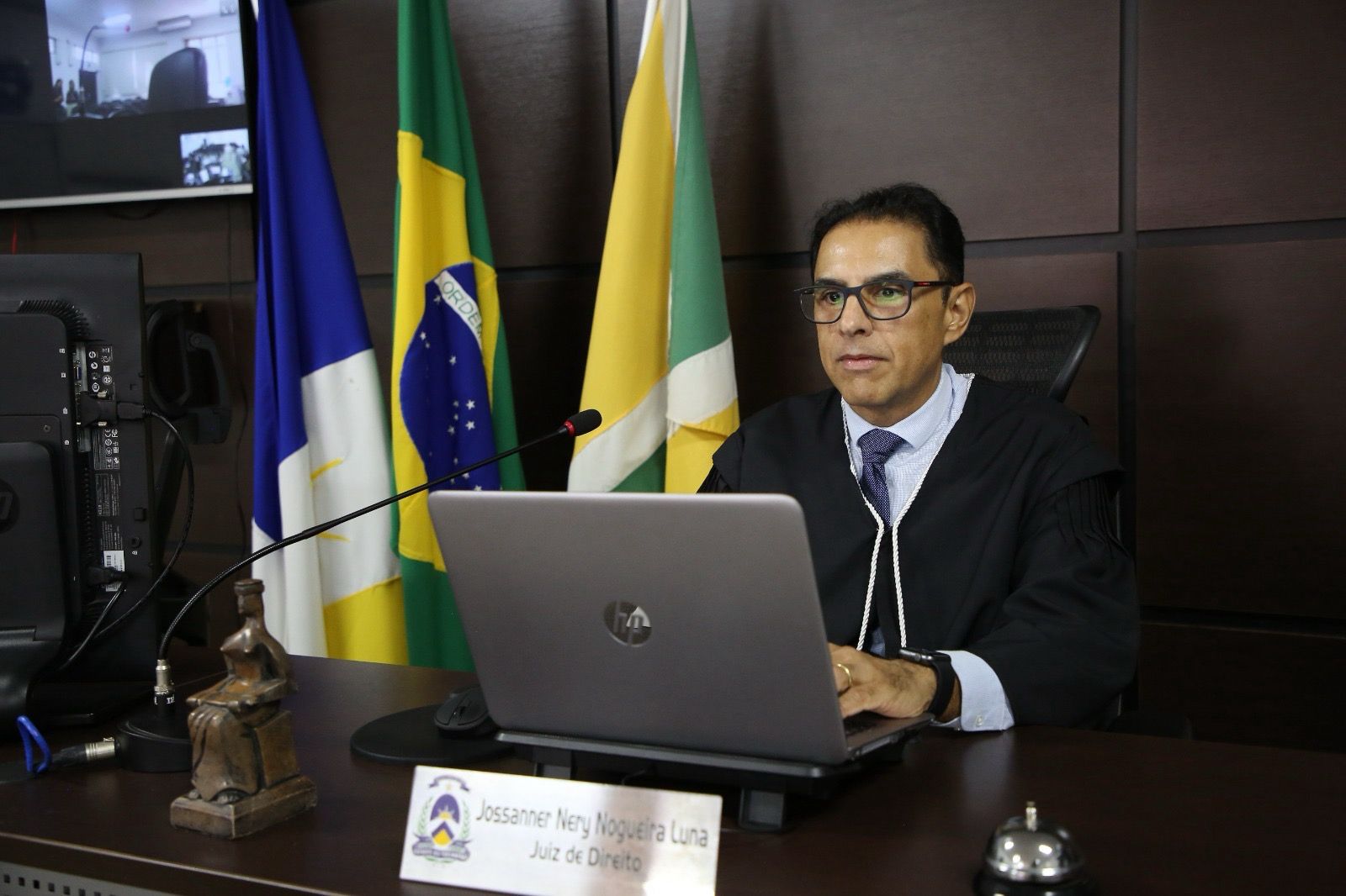 juiz Jossanner Nery, um homem moreno claro, usa óculos com armação escura; ele usa camisa branca e gravata e beca escura, está sentado, usando um notebook