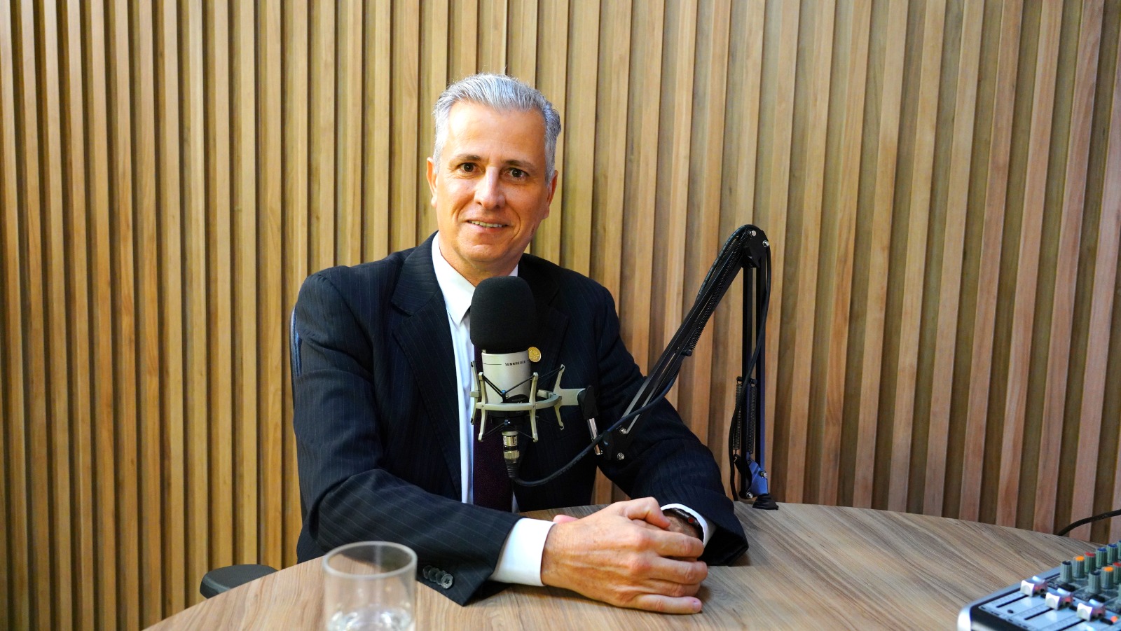 Fotografia colorida de um homem com terno, gravata e de cabelos grisalhos, sentado em uma cadeira, com os braços apoiados em uma mesa e com um microfone em sua frente. Ao fundo, uma parede de madeira listrada.