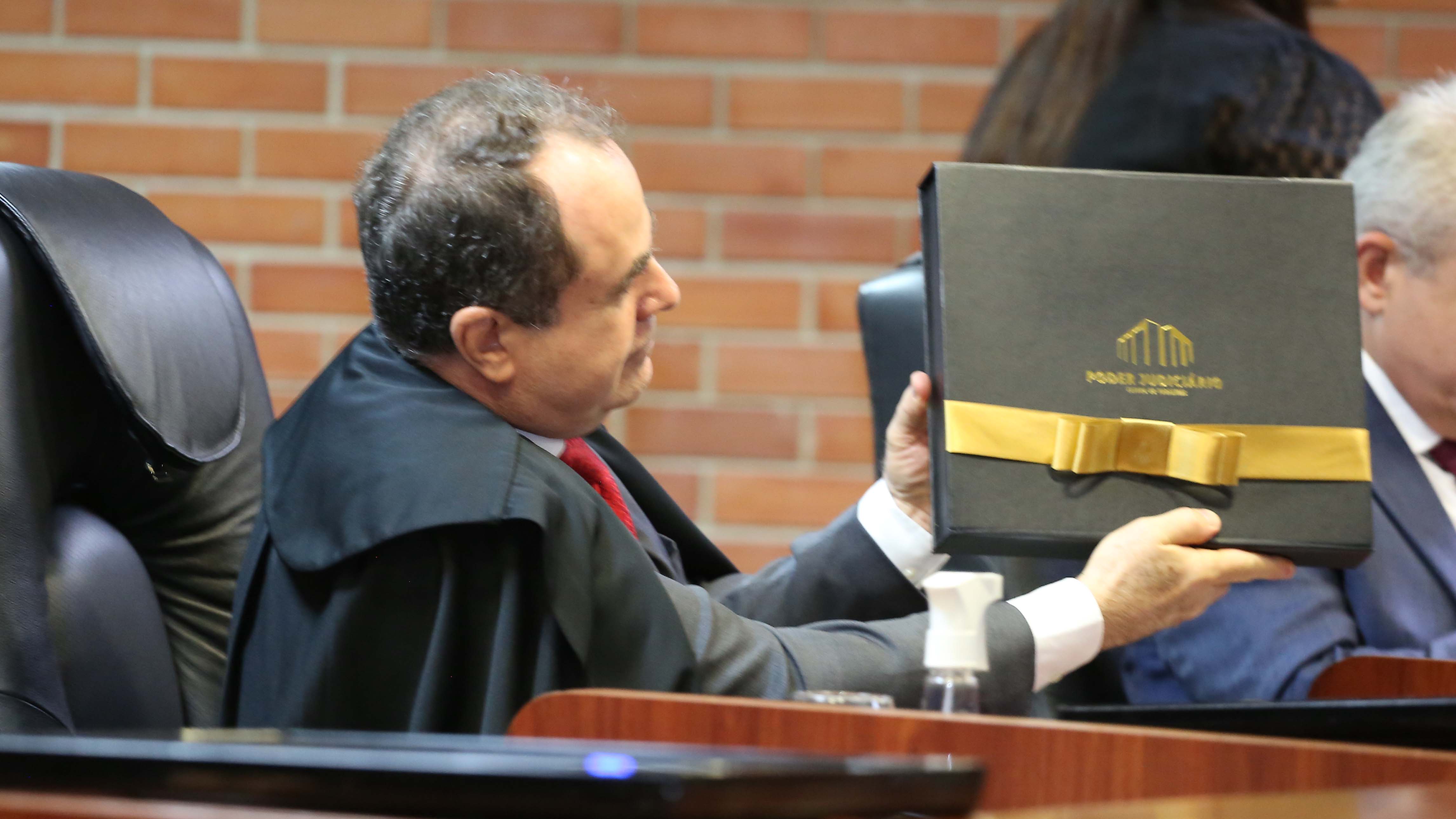 Imagem mostra um homem branco usando uma toga preta e segurando uma caixa preta com laço dourado
