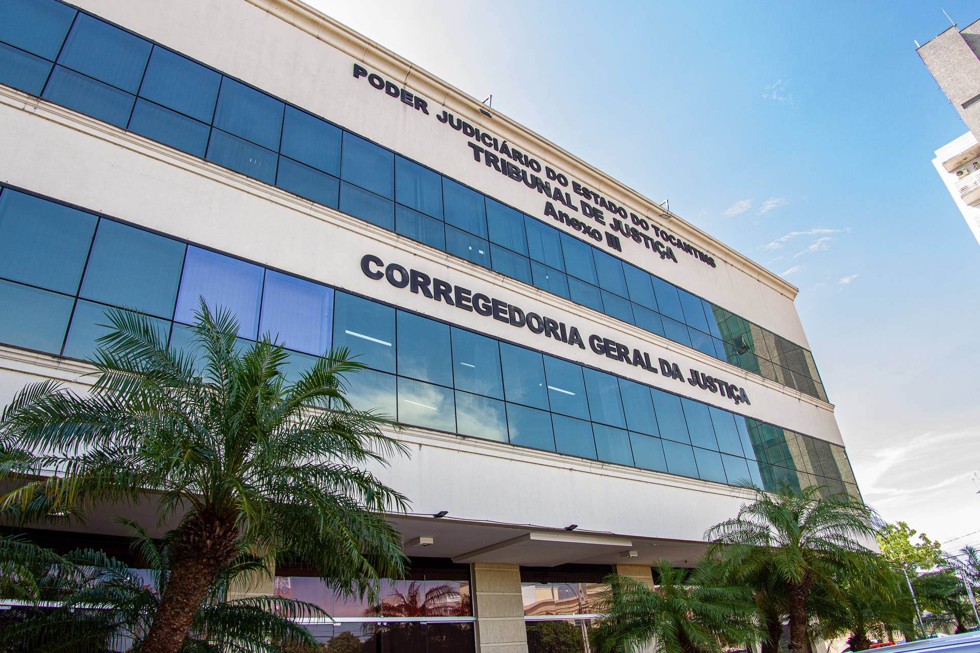 Fotografia colorida da fachada do prédio da corregedoria geral de justiça com algumas palmeiras na frente 