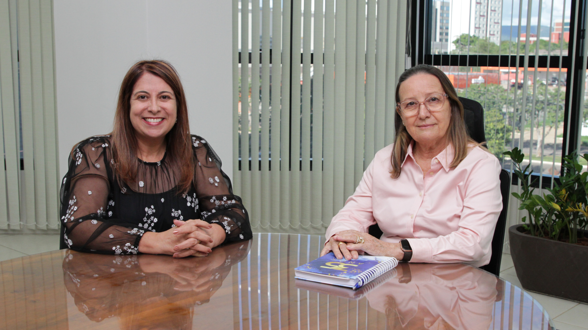 Fotografia colorida onde a Juíza Odete Almeida está sentada ao lado da  desembargadora Maysa Vendramini Rosal, de dedos laçados e apoiada sobre a mesa com a agenda dos 35 anos do Tribunal de Justiça do Tocantins em sua frente 