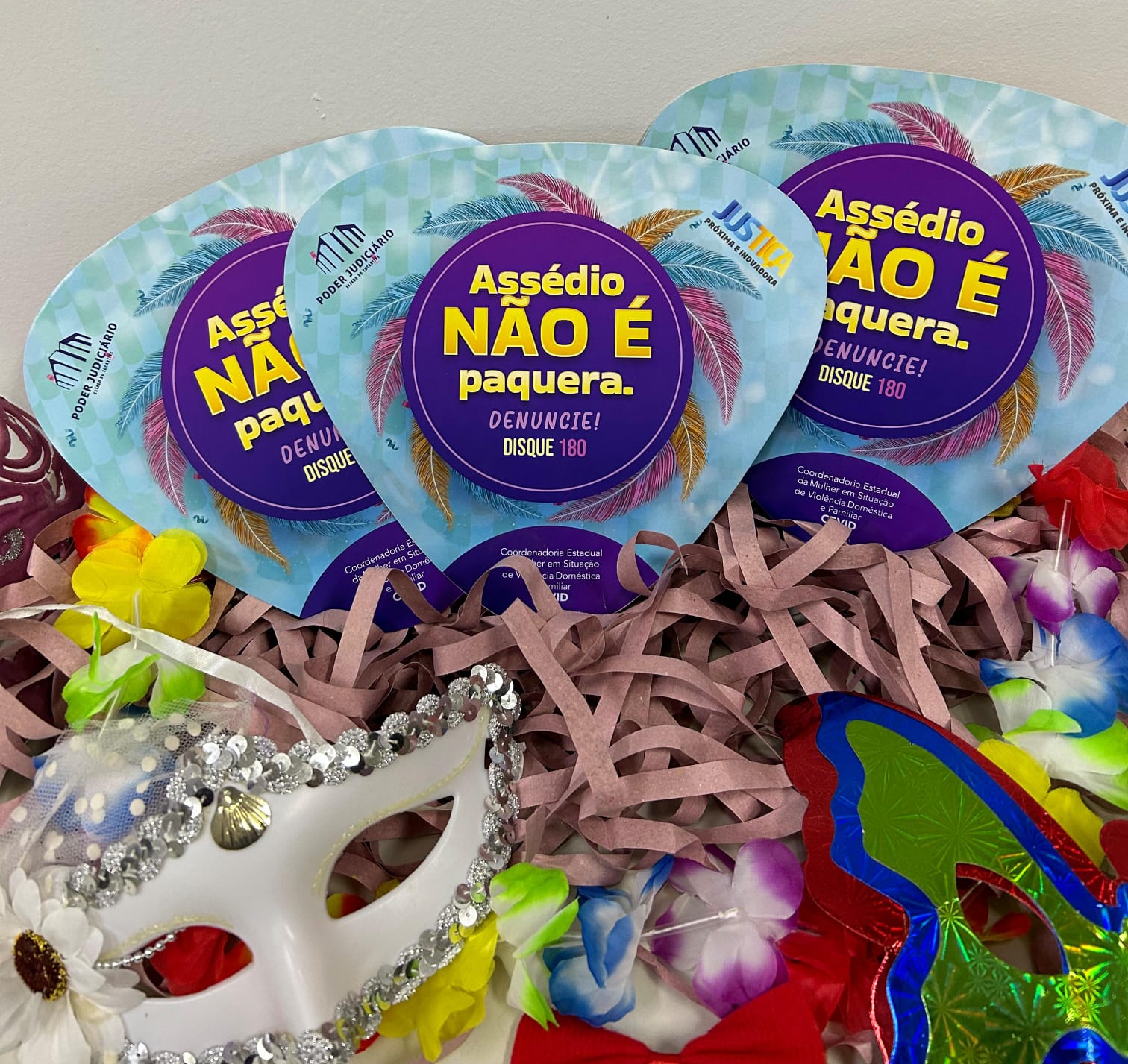 Fotografia colorida dos leques comemorativos da campanha "Assédio Não é Paquera", em cima de vários fios de papel bege, com duas máscaras ao lado, uma branca e outra colorida, e um colar de flores em volta 
