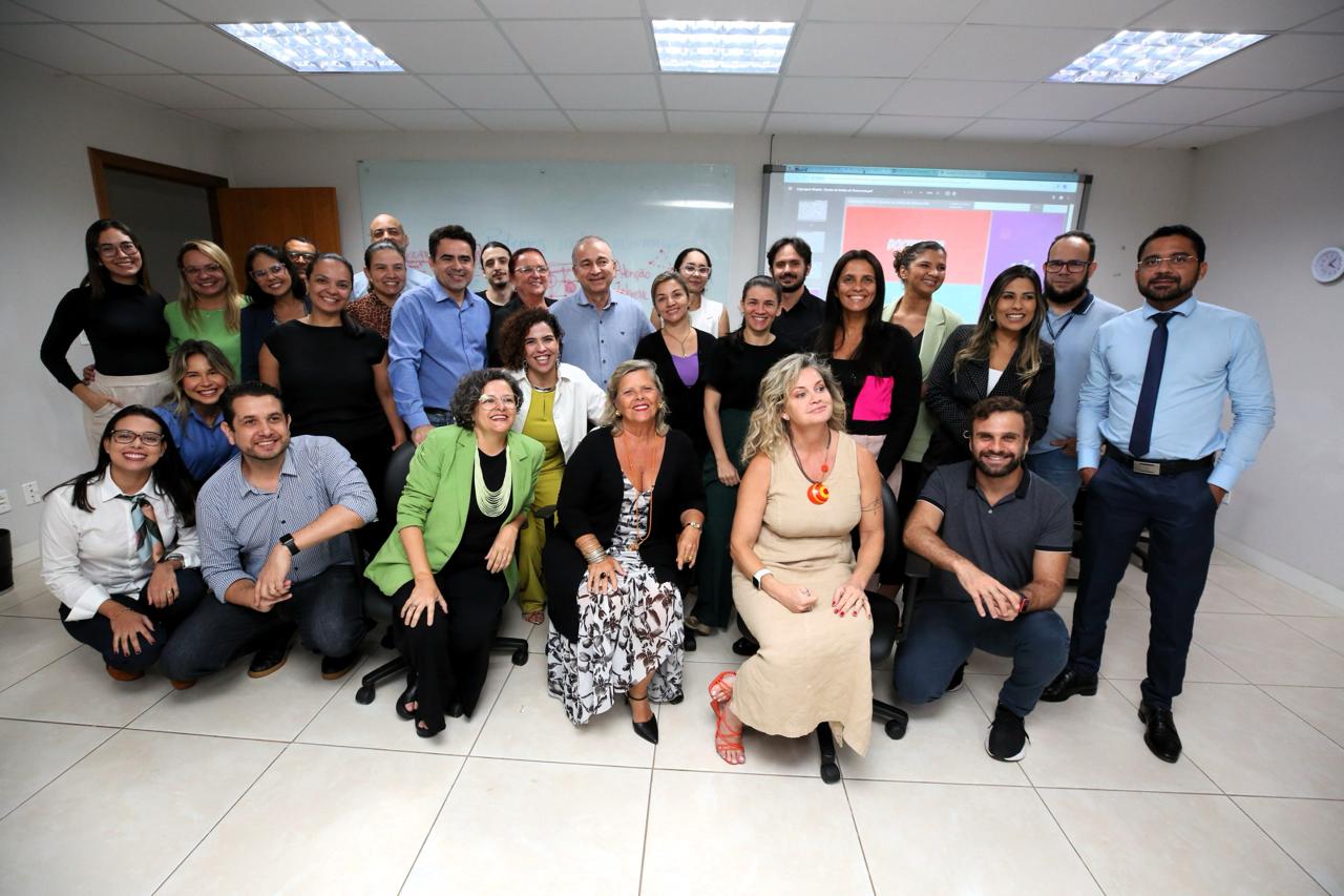 Fotografia colorida com homens e mulheres posando sentados e em pé, numa sala de aula 