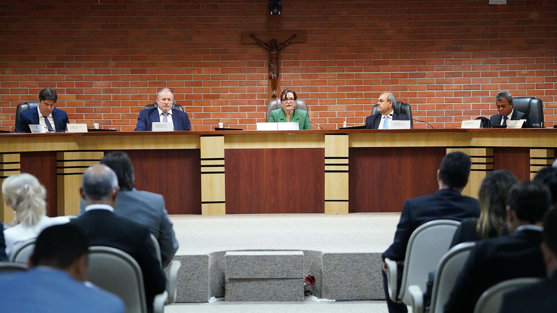 Imagem mostra pessoas sentadas em cadeiras; à frente uma mesa de madeira oval e à frente uma plateia