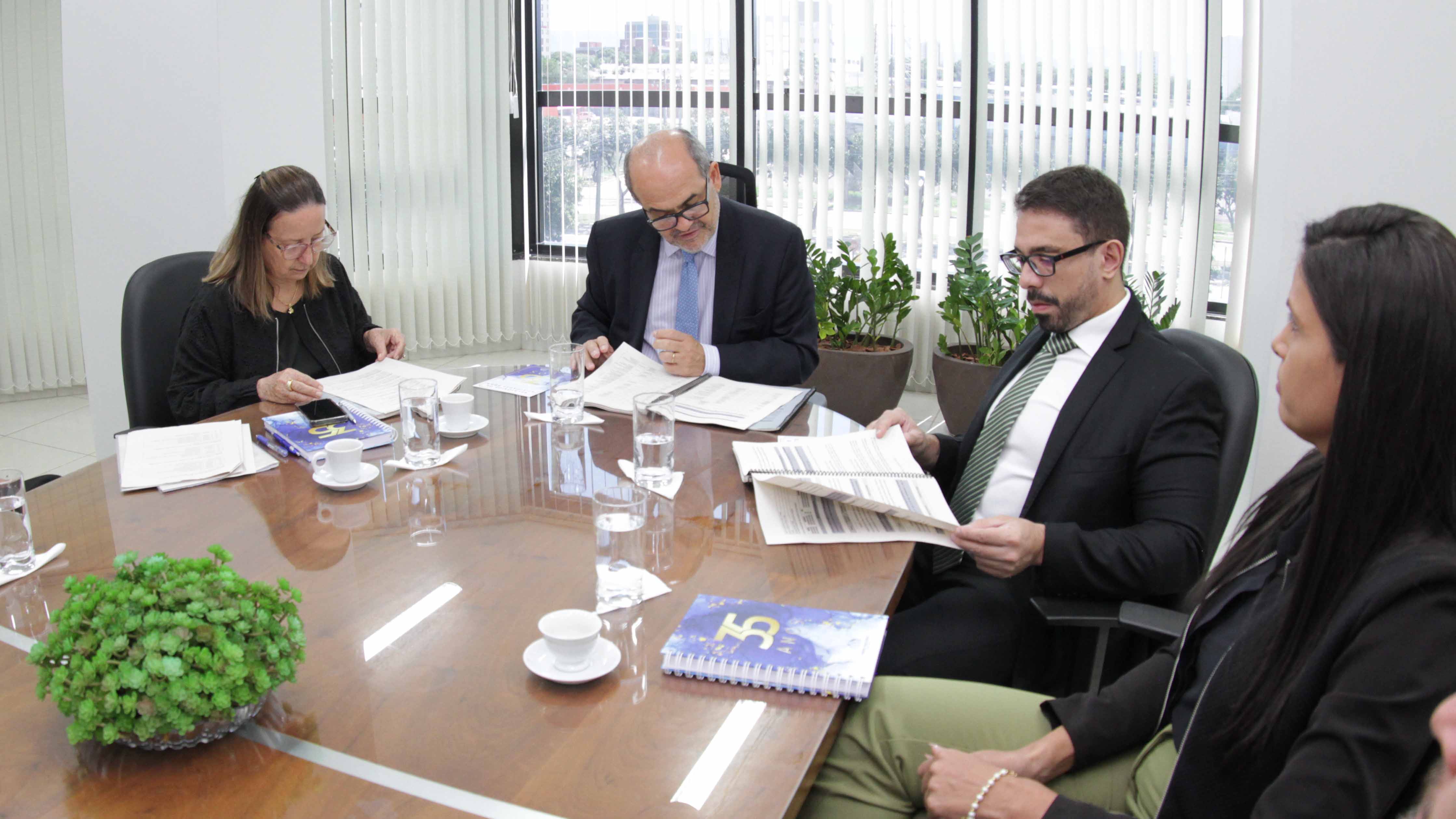Homens e mulheres vestidos de preto analisam documentos em mesa de reunião em sala branca com janela com persiana vertical aberta