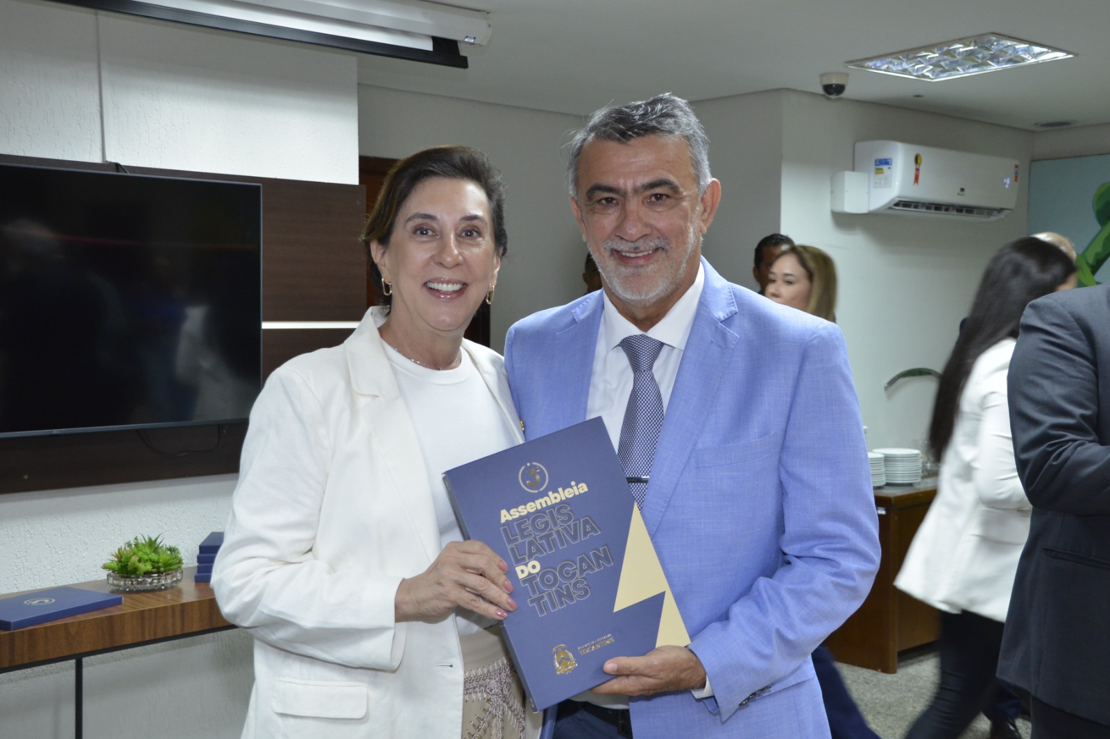 Imagem mostra um homens de terno azul e uma mulher de branco, em pé, segurando um livro