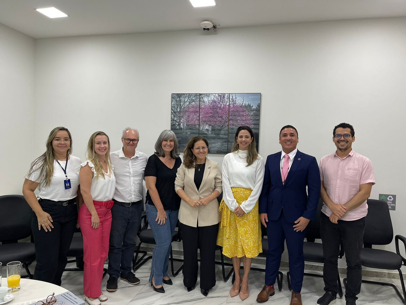 Homens e mulheres em pé em foto posada. Ao fundo, parede branca com quadro com imagem de natureza