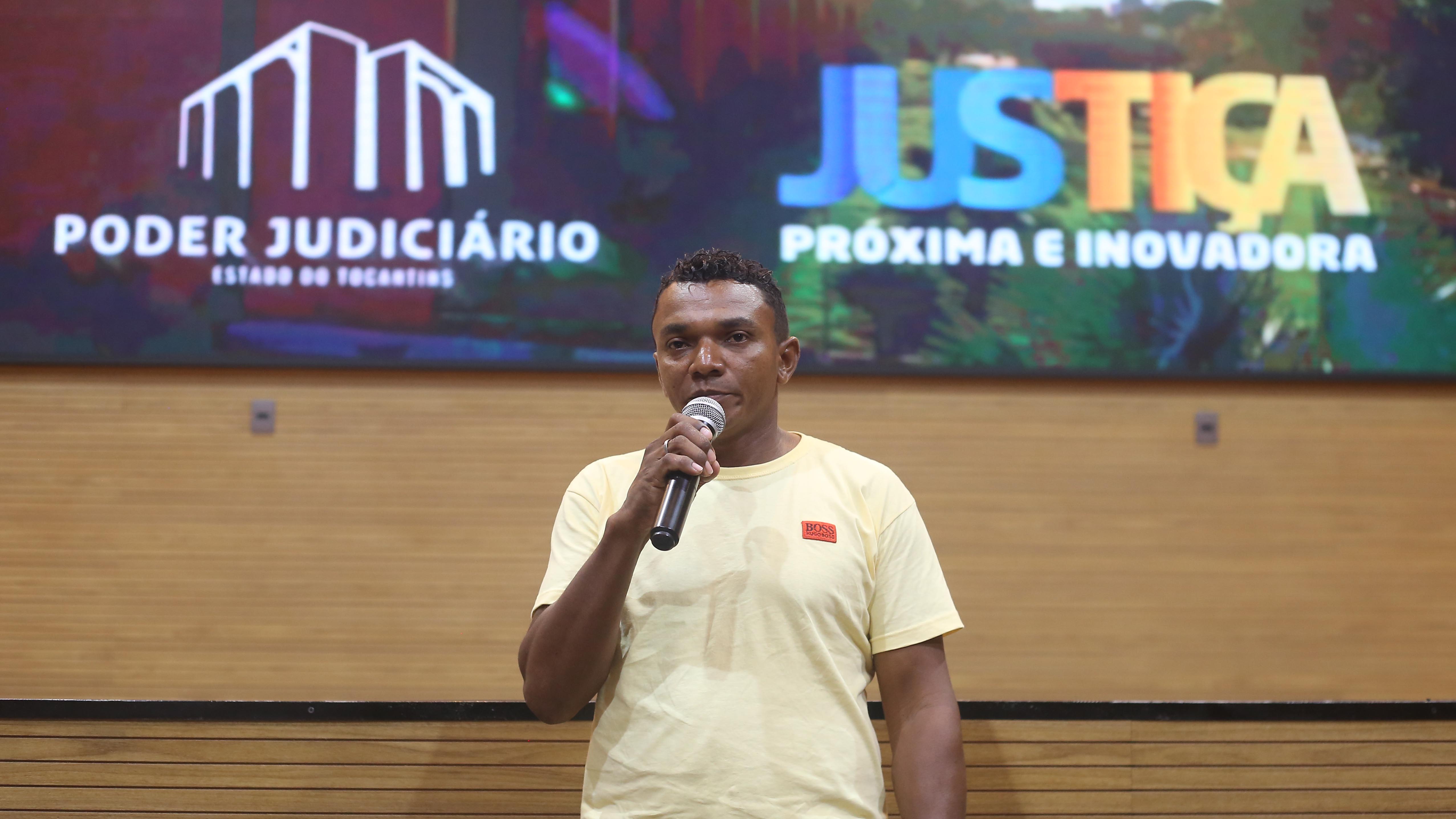 Homem negro de camiseta amarela fala ao microfone para plateia em auditório