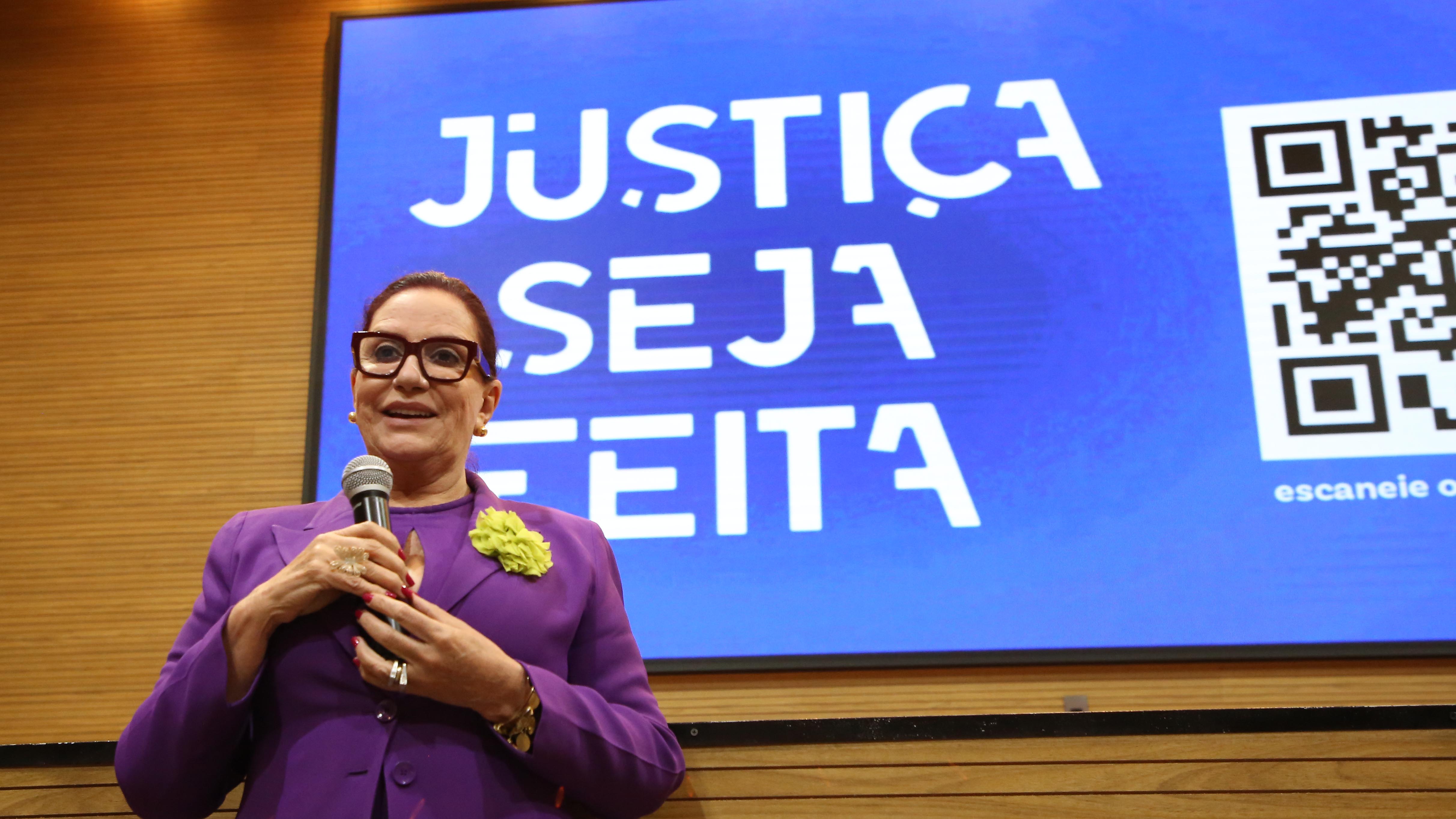 Fotografia colorida com uma mulher branca, usando lilás e óculos de grau, falando ao microfone