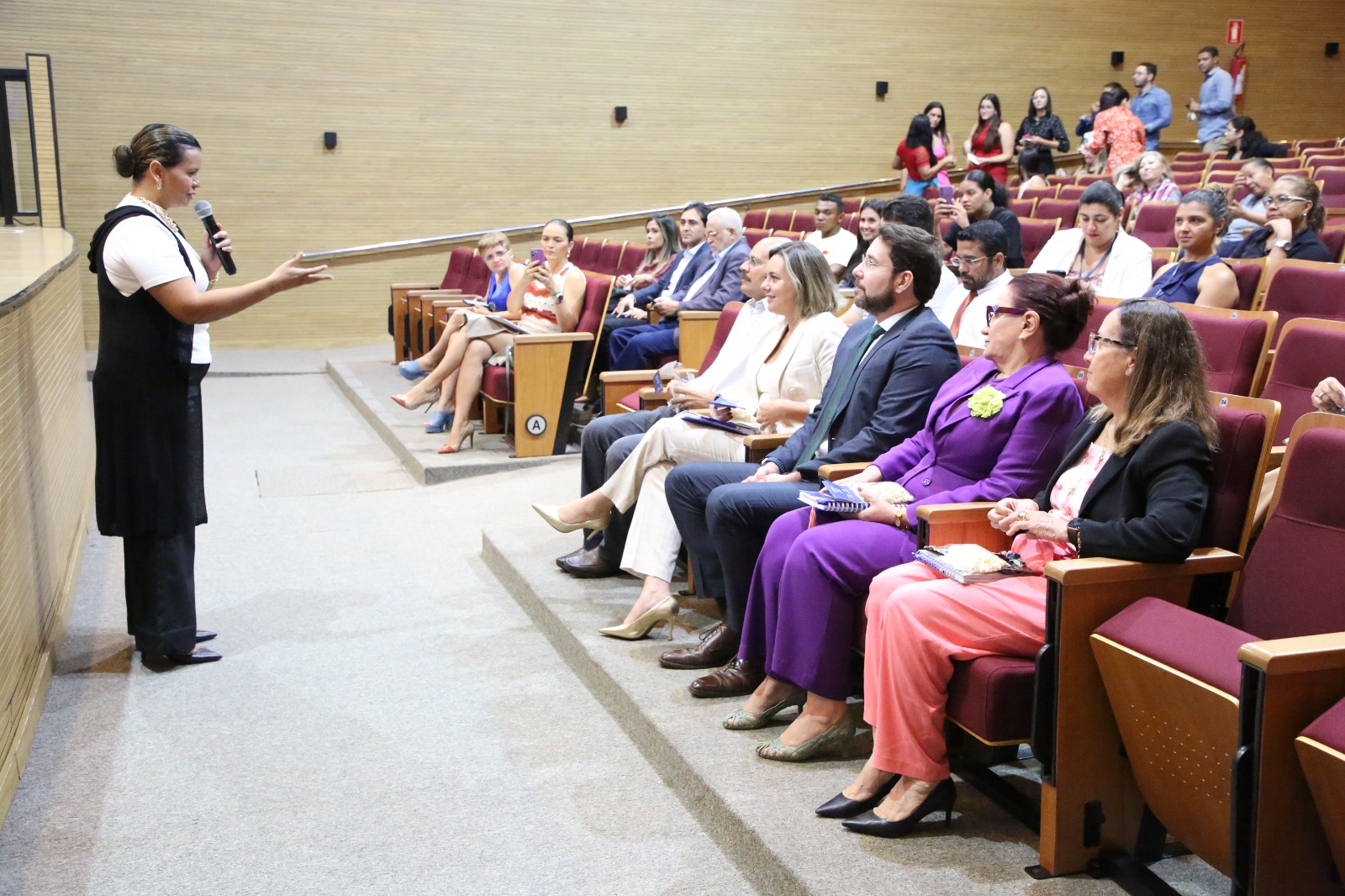 Fotografia colorida que mostra uma mulher parda, usando roupa preta e branca, falando para a plateia num auditório 