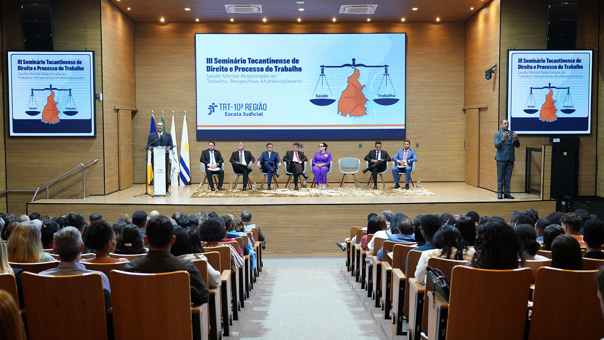 auditorio cheio com pessoas sentadas na plateia e no palco sete pessoas sentadas em cadeiras enquanto uma de pé fala ao microfone