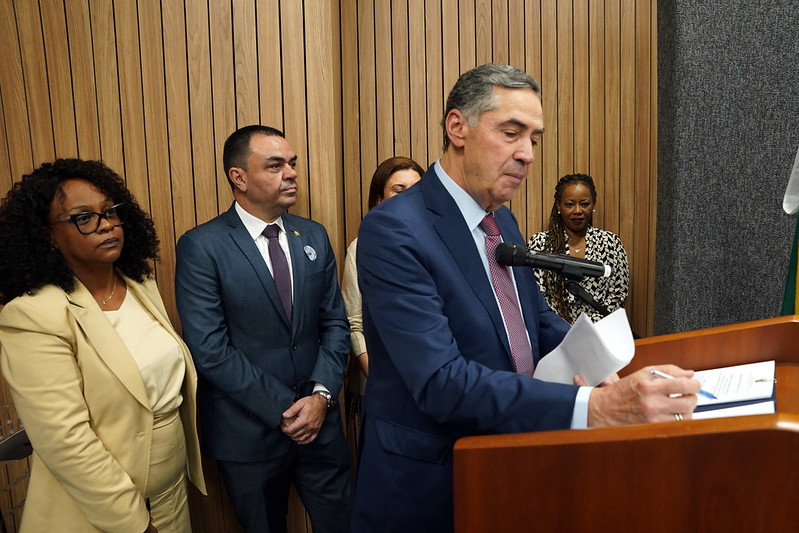 Fotografia colorida de cinco pessoas, dois homens brancos, uma mulher branca e duas mulheres negras. Um dos homens brancos, o ministro Luís Roberto Barrosoo, está falando sobre um palanque de madeira enquanto lê documentos.