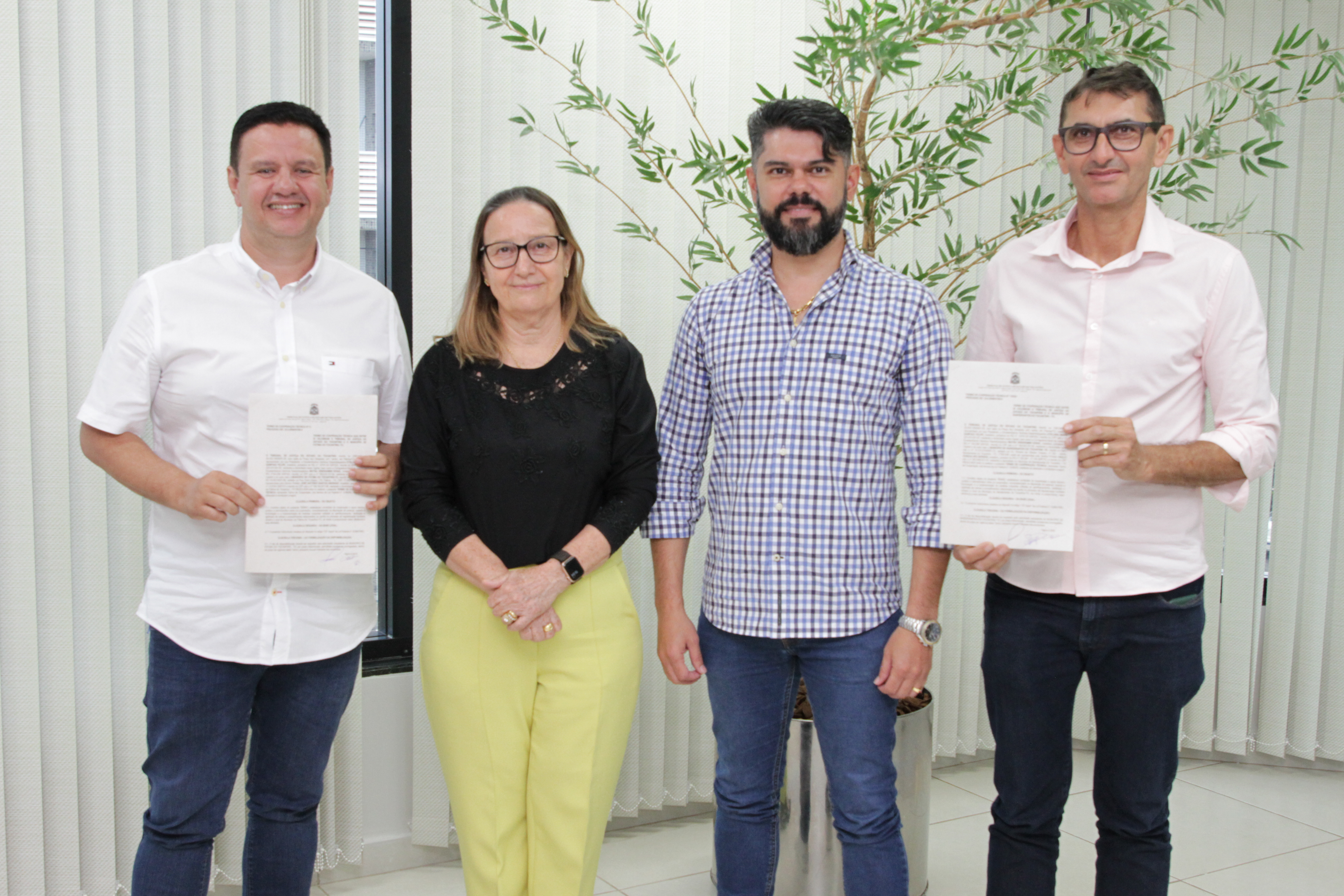 Fotografia colorida de três homens, vestidos de calça e camisa aberta de botão, e uma mulher, de calça amarela e camisa preta. Os homens de cada ponta estão segurando documentos de regularização fundiária 