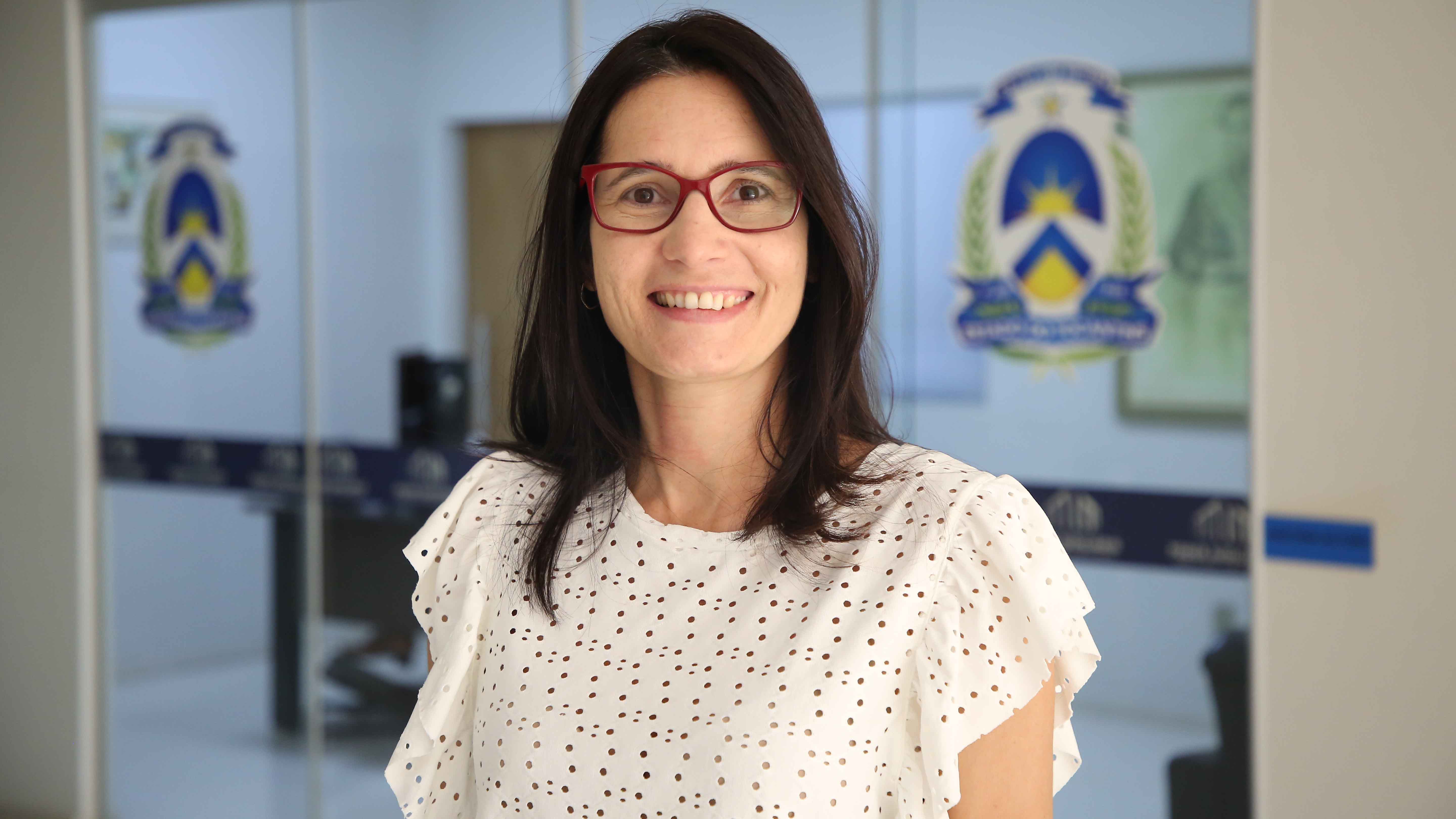 Marluce, responsável técnica do projeto; uma jovem de cabelos escuros, de óculos; ela usa uma blusa bege e ao fundo uma porta de vidro com a logo do Poder Judiciário