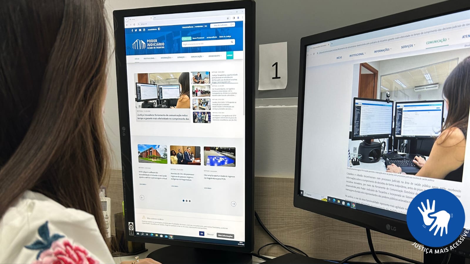 Fotografia colorida de uma jovem em frente a dois monitores de computador, um em pé e outro deitado, lendo uma matéria do site do Tribunal de Justiça do Tocantins em um dos monitores e com o portal aberto na parte principal em outro