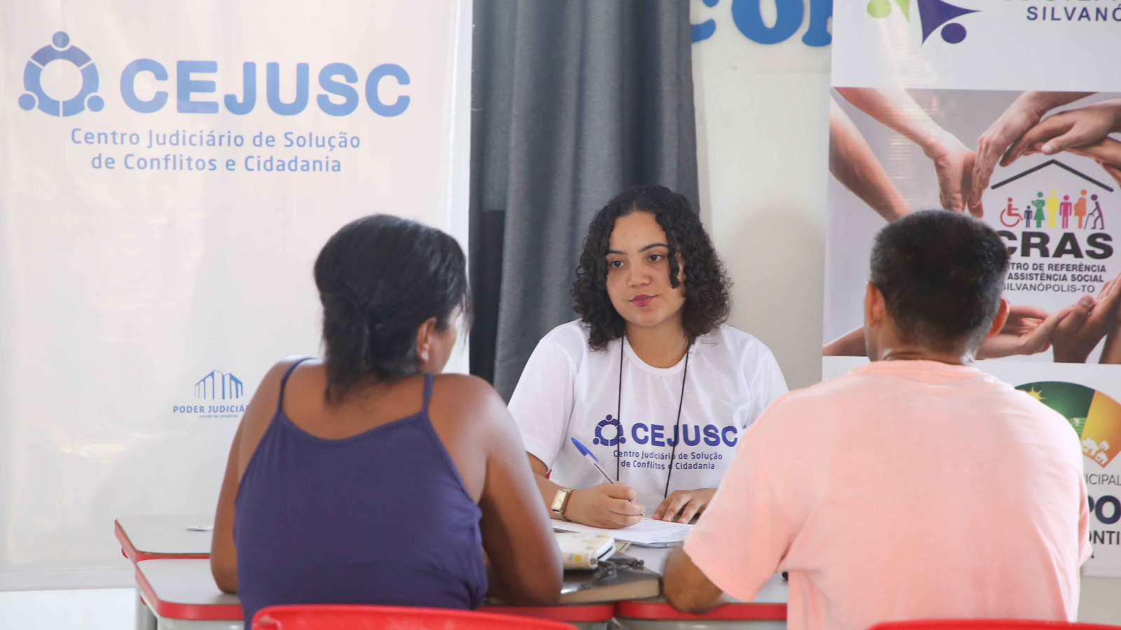 Casal sentado de costas em atendimento no Cejusc; de frente uma conciliadora sentada  vestida numa camiseta branca com o nome Cejusc