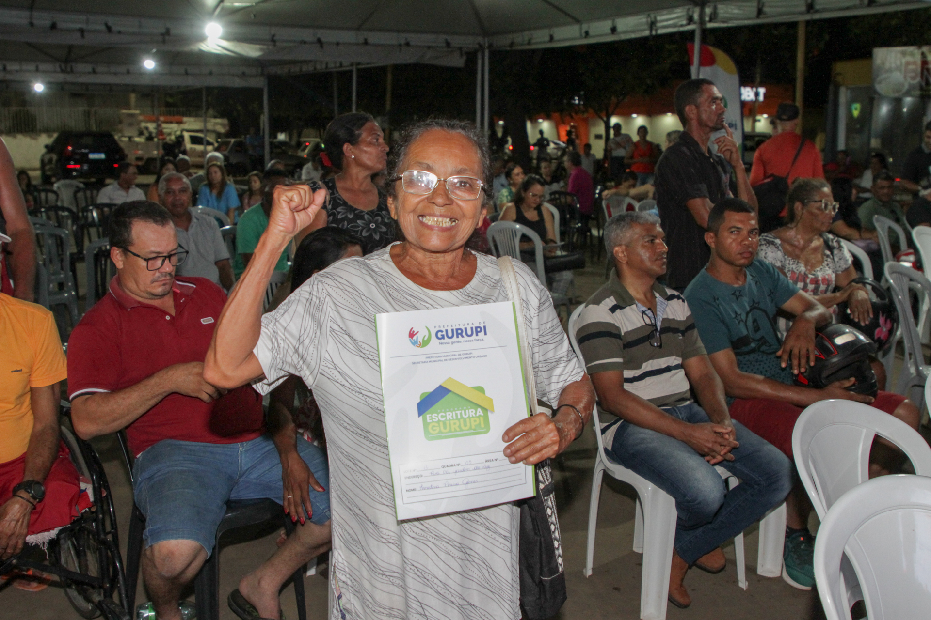 Fotografia colorida de senhora de vestido branco listrado com cinza claro e óculos segurando um documento de regularização fundiária enquanto pessoas estão sentadas atrás dela em cadeiras de plástico 