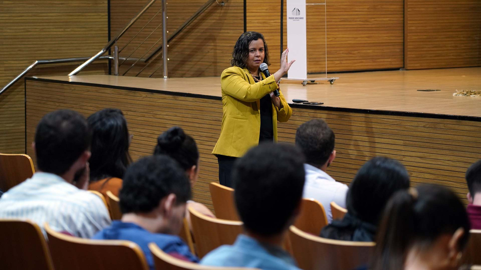 Kézia, ministrando palestra, em pé, fala ao microfone, usa roupa preta e blazer mostarda; servidores de costas, assistindo a palestra