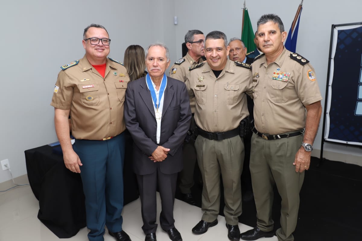 juiz aposentado Rosemilto ao centro, de terno escuro e camisa branca, com uma medalha no pescoço; ele segura uma pasta azul; ao seu lado, militares uniformizados e dois homens de camisa branca e calça escura