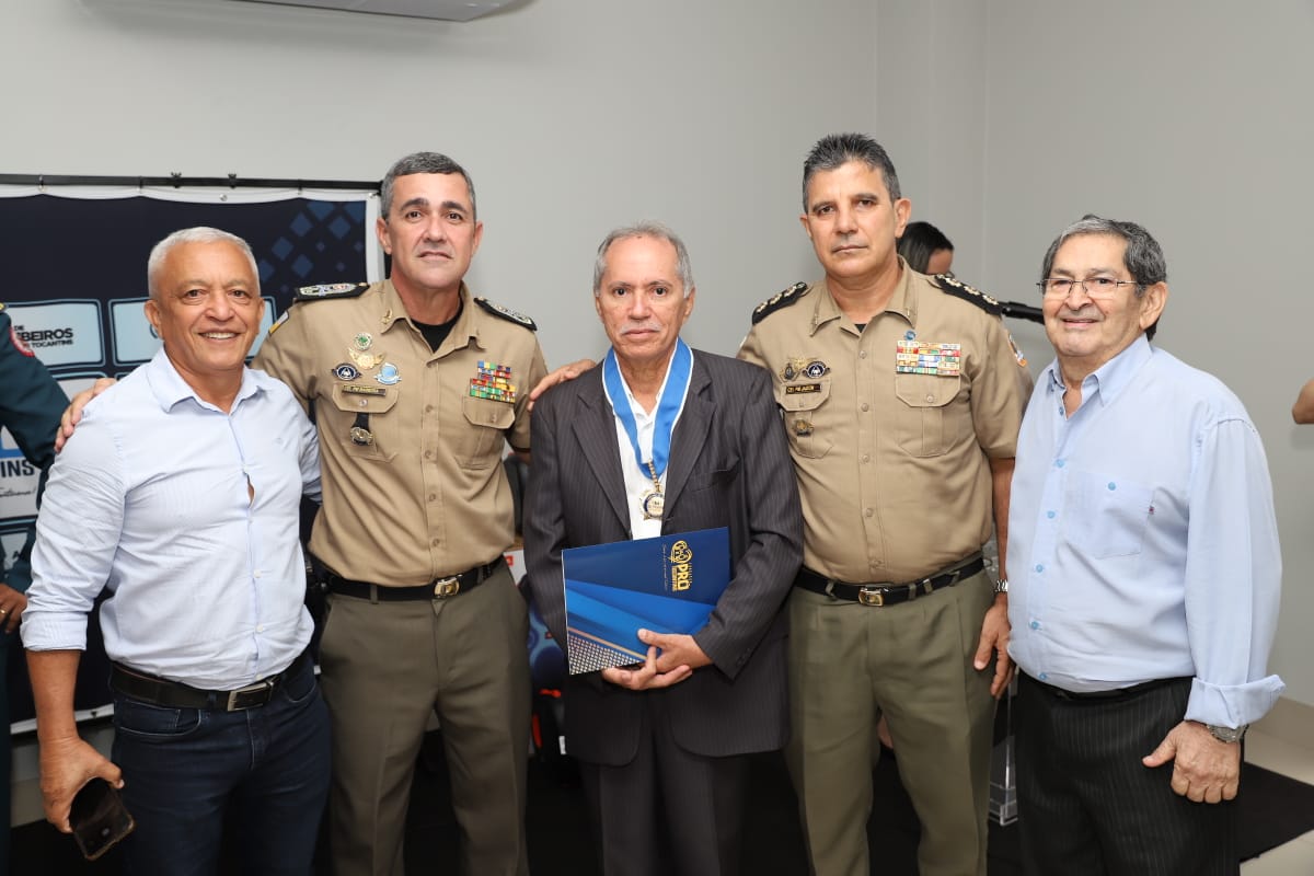 juiz aposentado Rosemilto ao centro, de terno escuro e camisa branca, com uma medalha no pescoço; ele segura uma pasta azul; ao seu lado, militares uniformizados e dois homens de camisa branca e calça escura