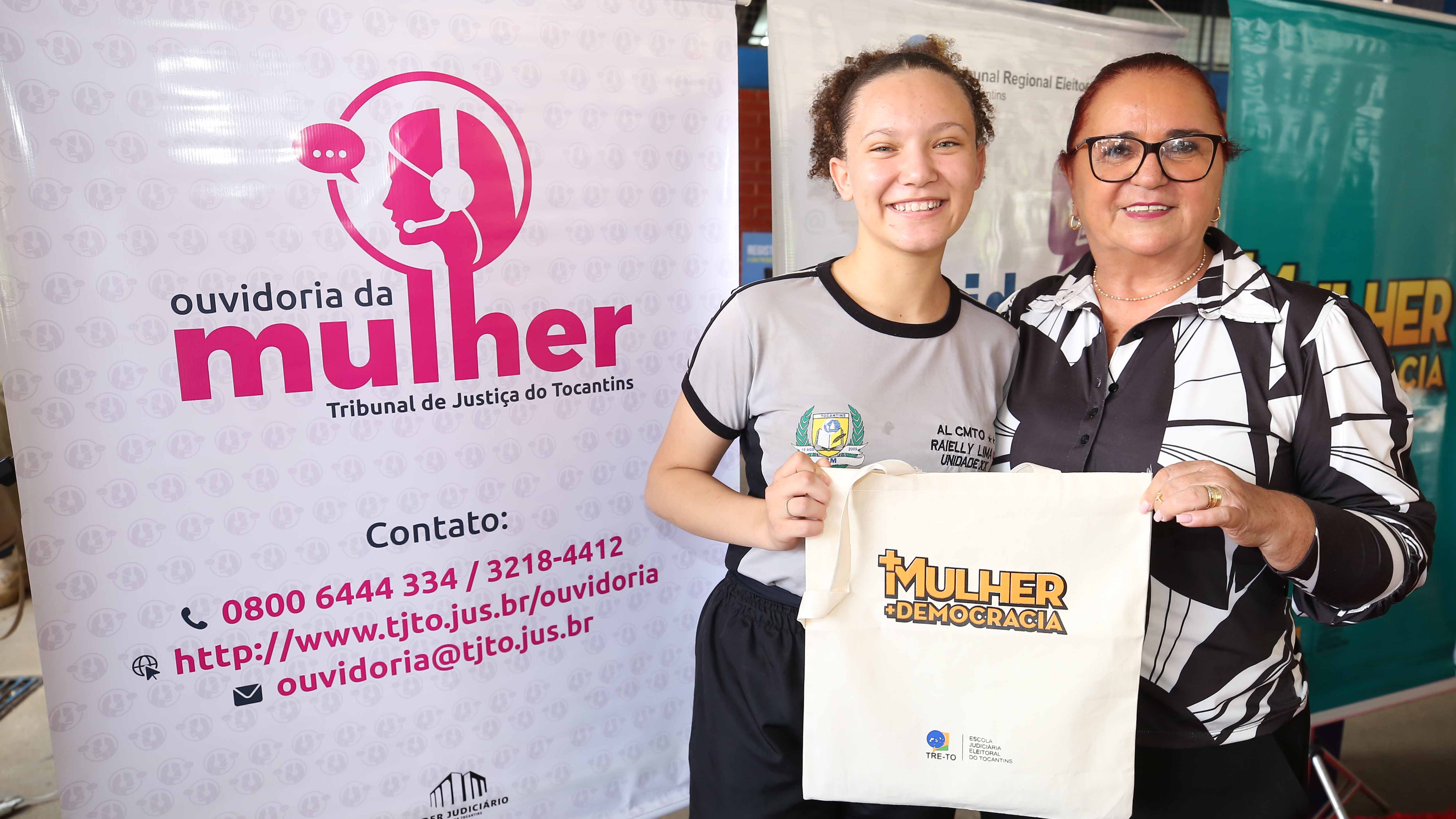 Aluna branca de cabelos amarrados e mulher parda, de óculos de grau, também de cabelos amarrados, seguram camiseta com estampa sobre projeto voltado para Mulher, ao lado de banner sobre Mulher