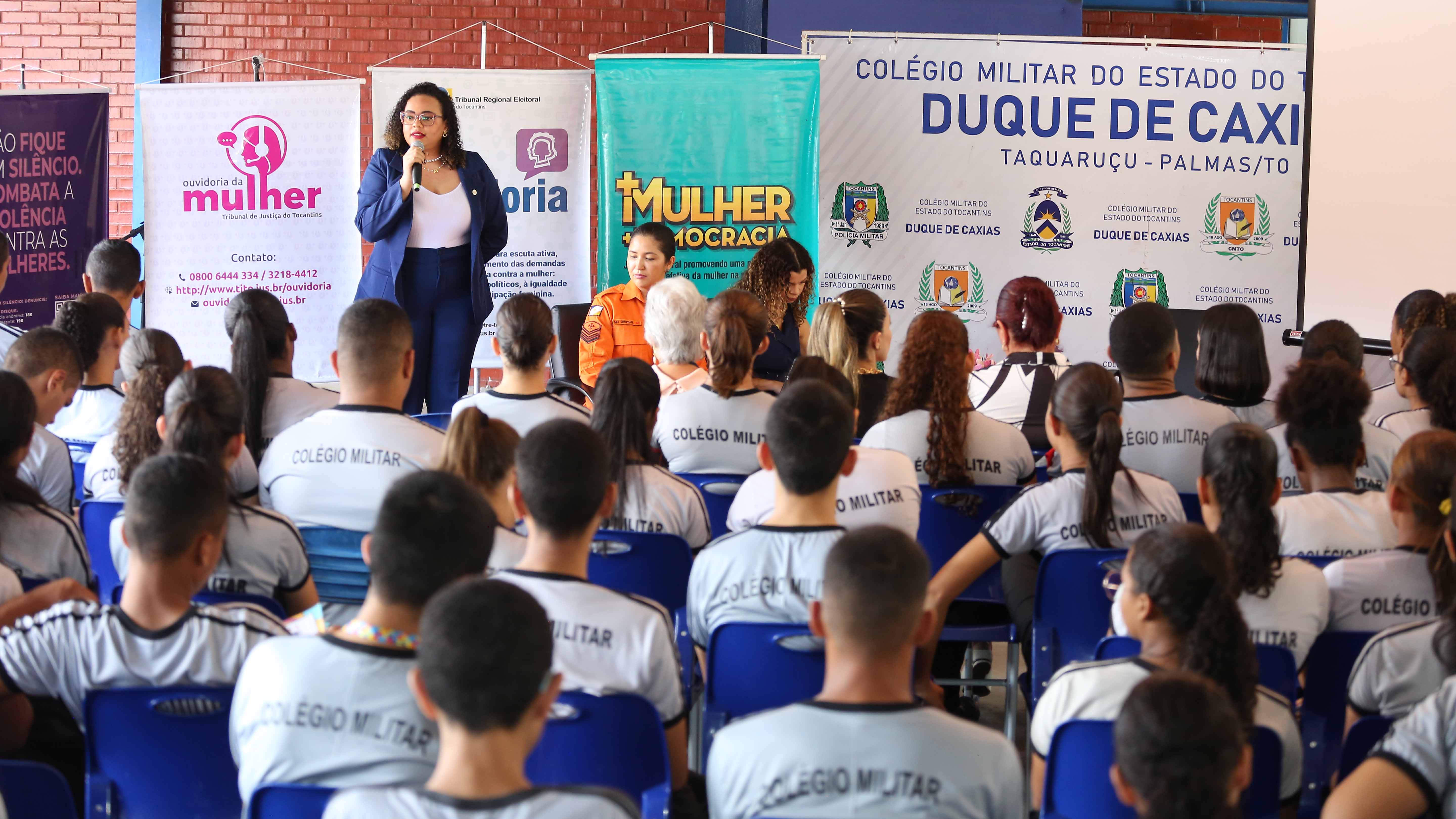 Alunos de escola sentados de costas assistindo palestra de mulher parda, cabelos cacheados soltos, vestida de terninho azul e camisa branca. Ao fundo, vários banners com informações sobre mulher