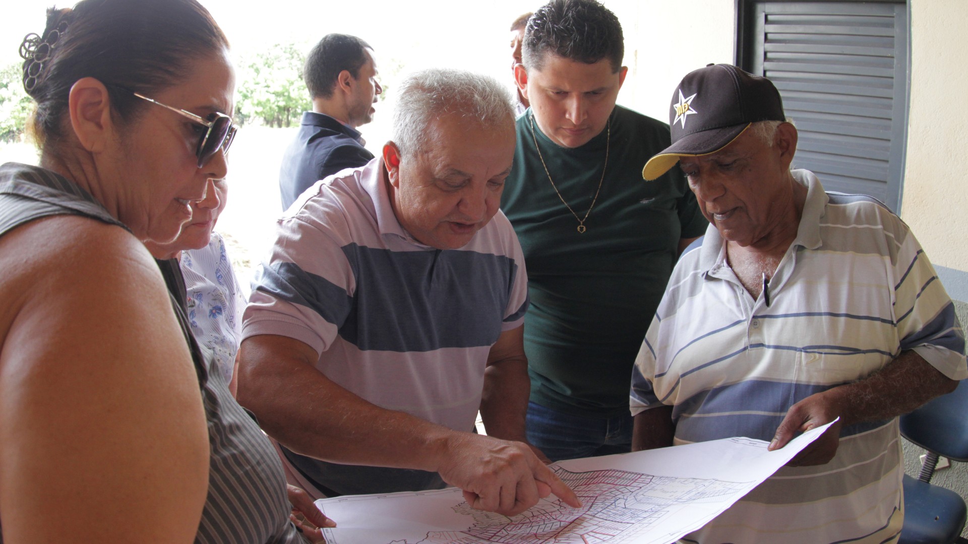 Fotografia colorida onde dois senhores (um deles de boné), três homens e uma mulher de óculos escuros olham uma planta de um terreno enquanto um dos senhores aponta um local na planta