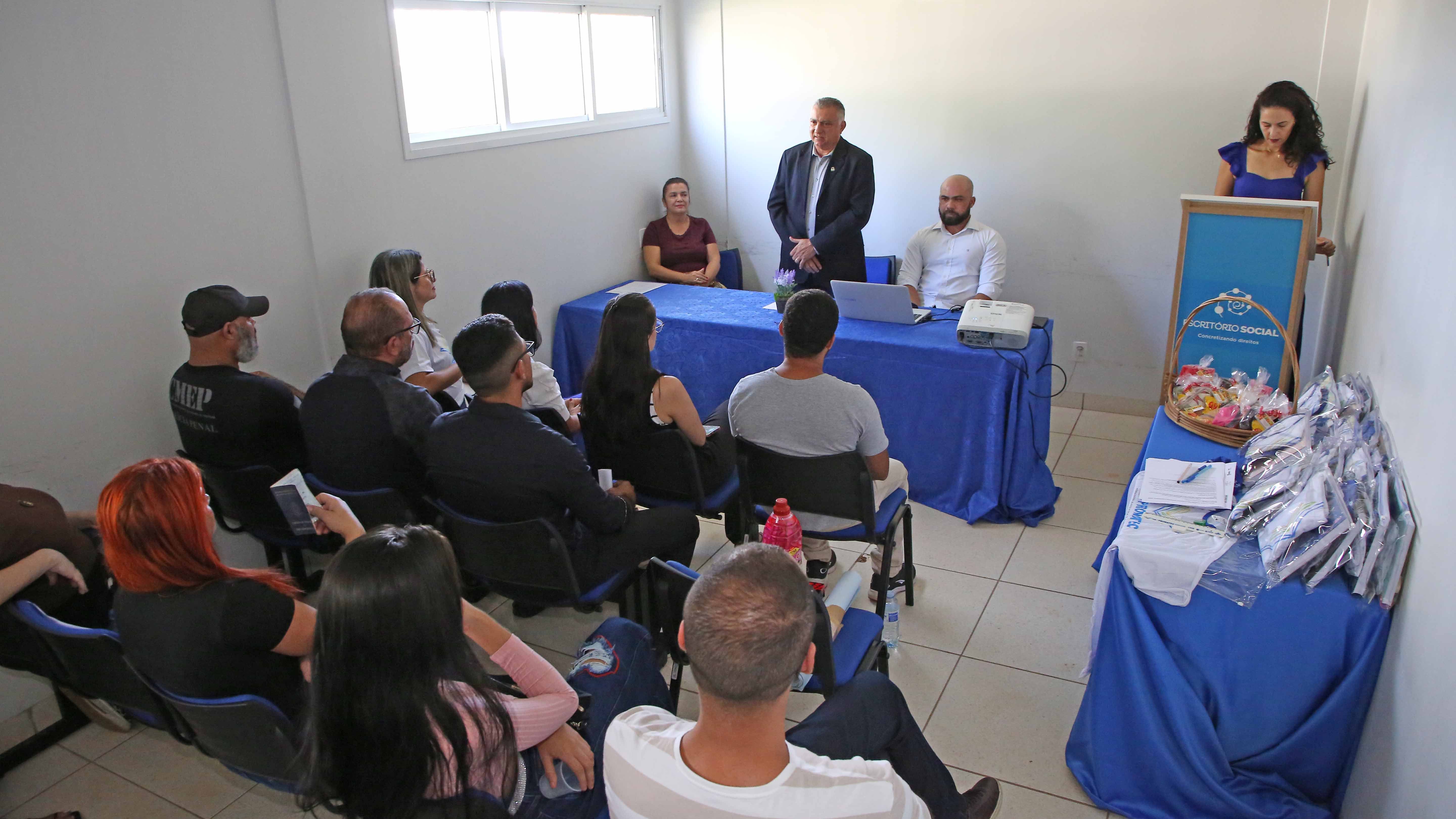 Homens e mulheres sentados em sala de paredes brancas assistindo aula. Duas mesas com tecido azul, uma na lateral com kits de caderno e caneta e outra ao fundo com três pessoas, uma mulher de camisa vinho sentada, um homem de camisa branca sentado e ao centro um homem de terno escuro em pé. Ao canto, púlpito em madeira com mulher de camisa azul e cabelos cacheados soltos. 