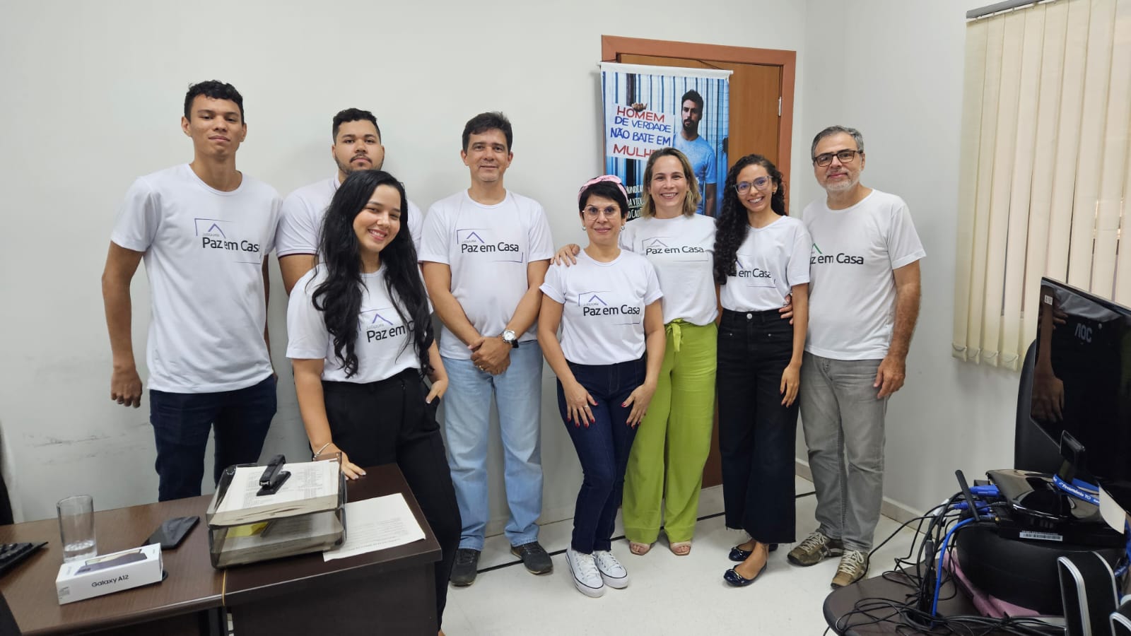 Homens e mulheres em pé em uma sala de paredes branca com camisa branca com estampa do Programa Justiça pela Paz em Casa 