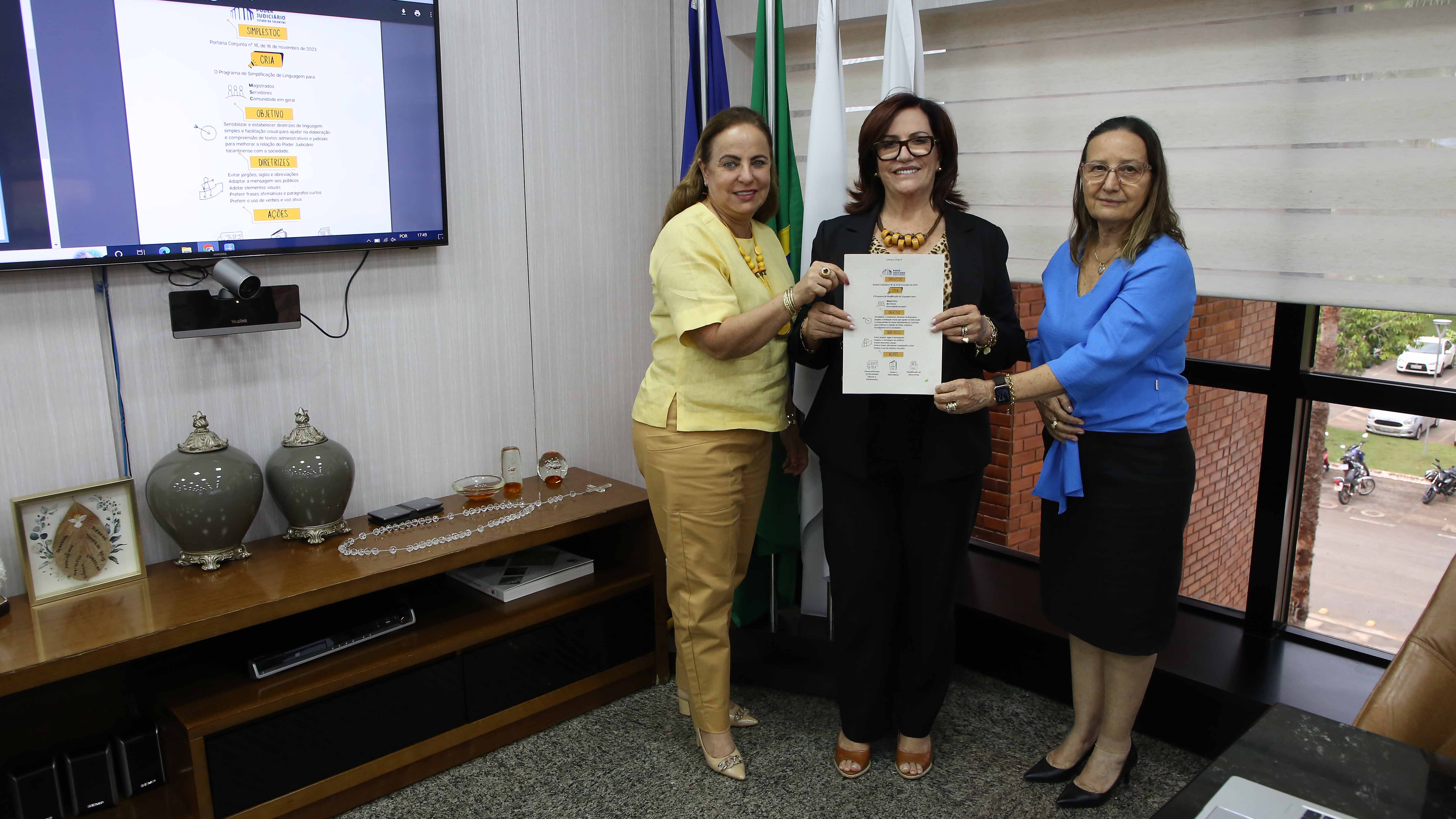 Imagem mostra três mulheres brancas, em pé, segurando um documento