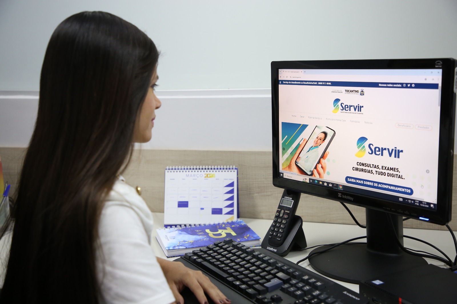 Mulher parda de cabelos lisos e longos sentada em frente a computador. Na mesa ainda tem agenda e calendário do Tribunal de Justiça e um telefone preto, a sala é branca