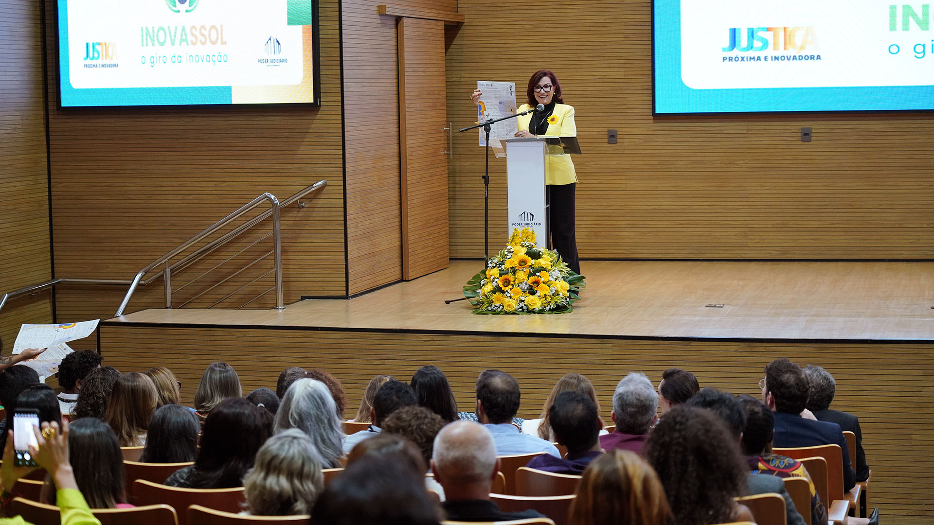 Imagem colorida de uma mulher branca de meia-idade usando roupa colorida (amarela e preta) e falando para um público 