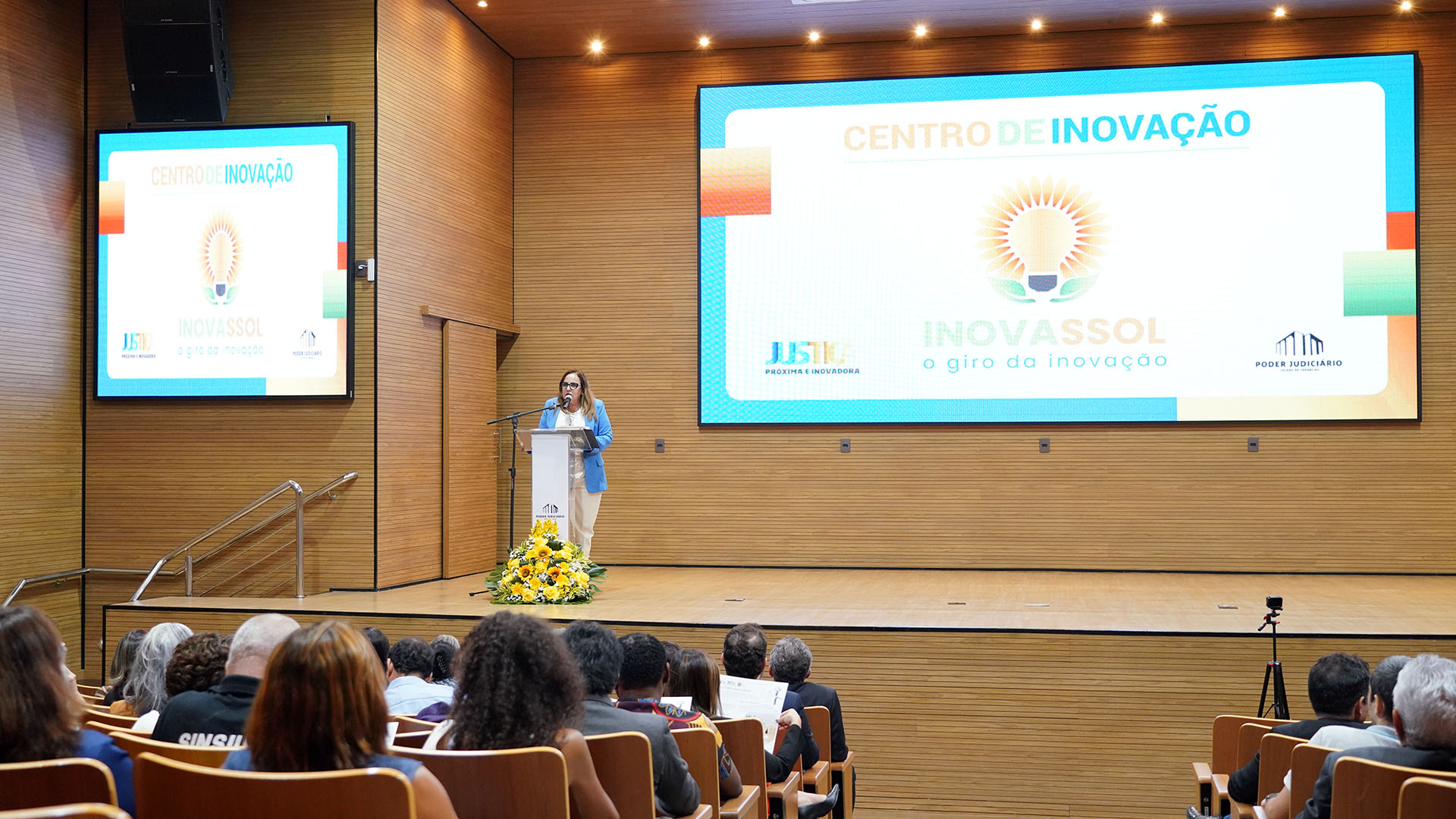 Imagem colorida de mulher branda de meia-idade vestindo roupa colorida (branca e azul) falando para a plateia 